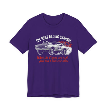 Purple Bella+Canvas 3001 T‑shirt with a bold graphic design. The print features a vintage muscle car with the number 69, white smoke and red flames, the distressed white title “The Meat Racing Channel” above, and the red script tagline “When the Steaks are high you can’t beat our meat” below. Funny drag racing parody shirt with retro hot rod style graphics.
