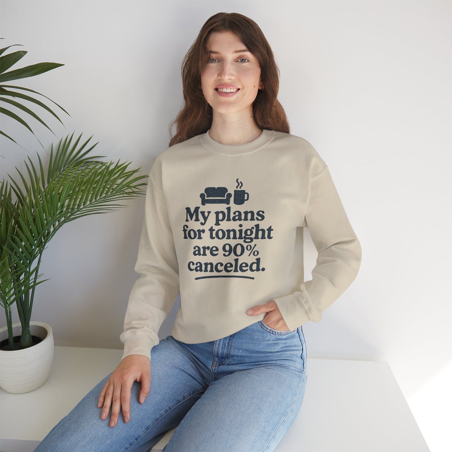 Woman wearing a beige sweatshirt with text 'My plans for tonight are 90% canceled' sitting on a white surface with a plant in the background