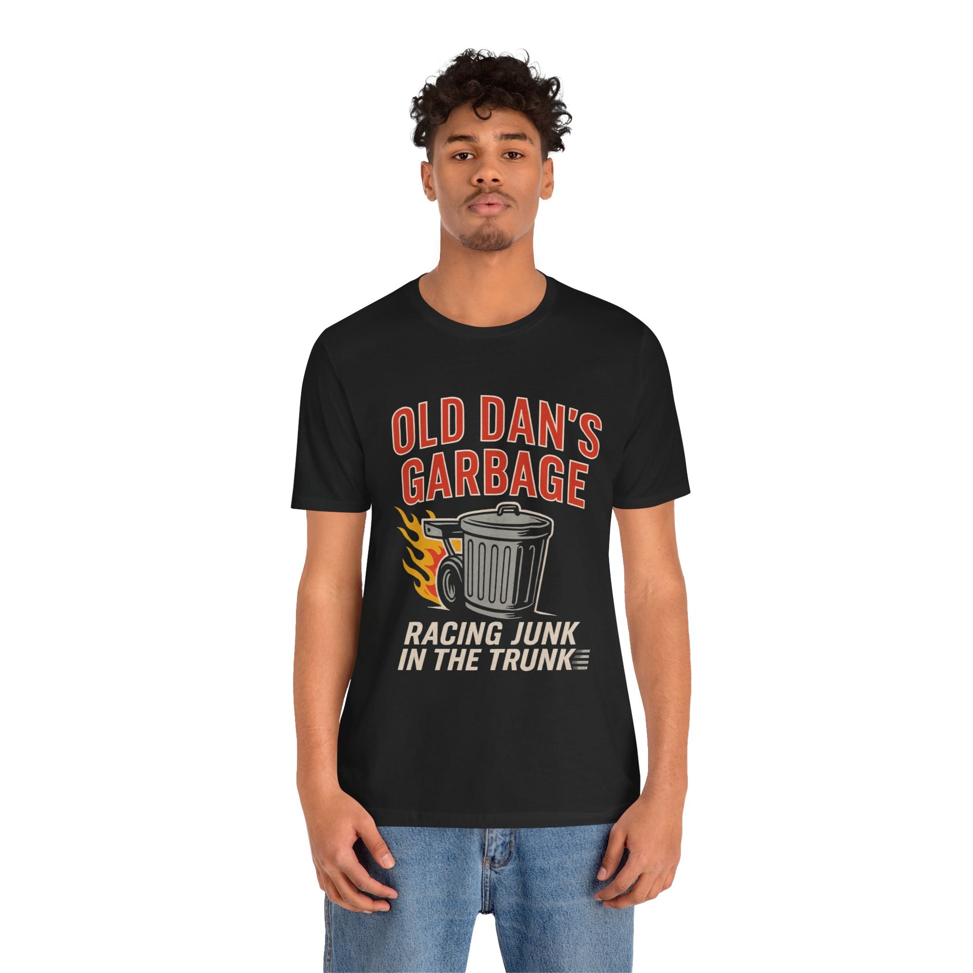 Person wearing a black graphic T‑shirt featuring the bold red and white text “Old Dan’s Garbage” above a flaming garbage can illustration, with the tagline “Racing Junk in the Trunk” below. Funny drag racing parody shirt with retro racing flames, checkered flag background, and humor‑driven hot rod style, perfect for car enthusiasts and gearhead apparel fans.