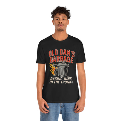 Person wearing a black graphic T‑shirt featuring the bold red and white text “Old Dan’s Garbage” above a flaming garbage can illustration, with the tagline “Racing Junk in the Trunk” below. Funny drag racing parody shirt with retro racing flames, checkered flag background, and humor‑driven hot rod style, perfect for car enthusiasts and gearhead apparel fans.