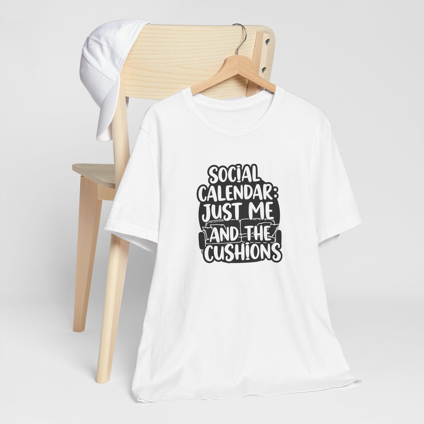 White t-shirt with text 'Social calendar: Just me and the cushions' design on a wooden chair against a white background