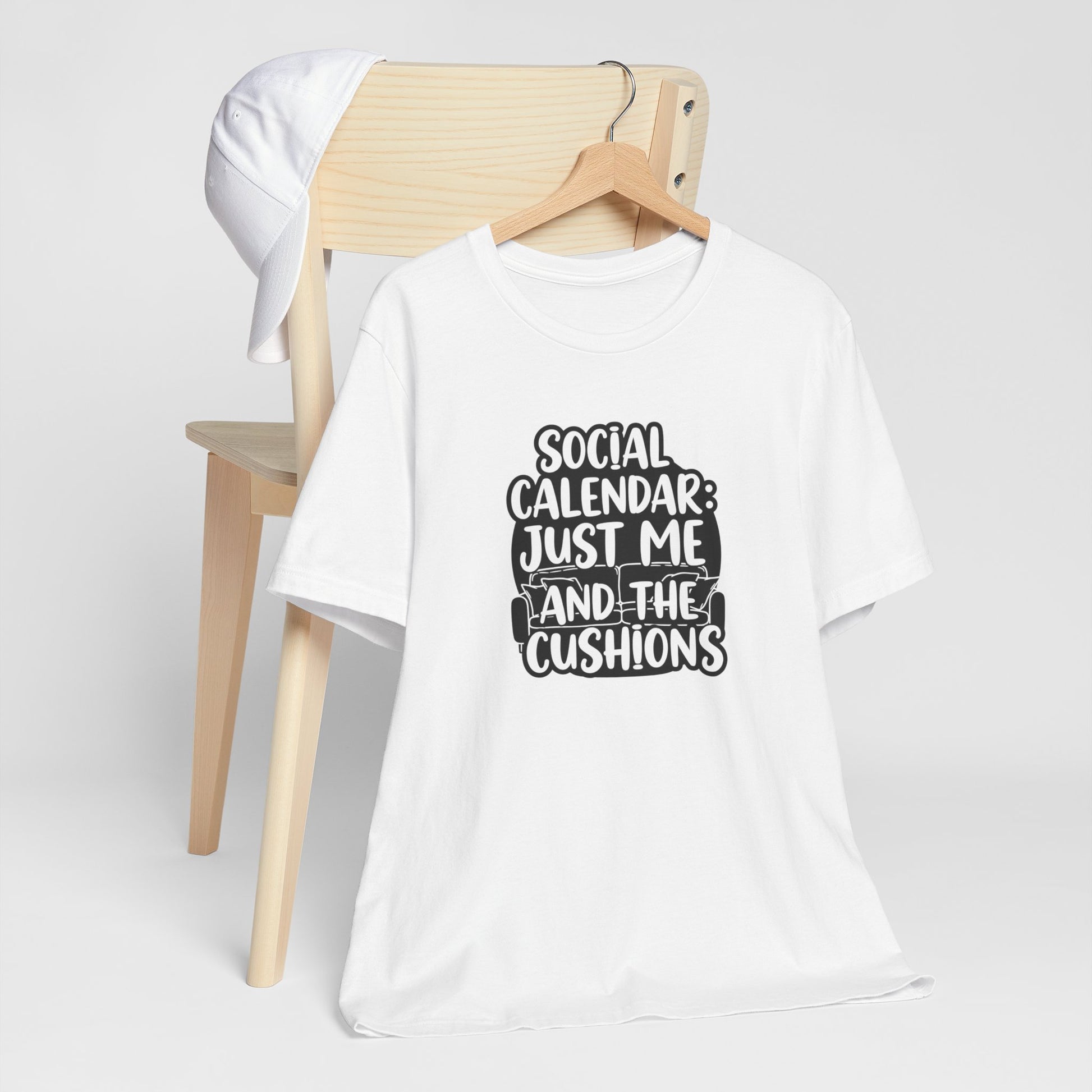 White t-shirt with text 'Social calendar: Just me and the cushions' design on a wooden chair against a white background