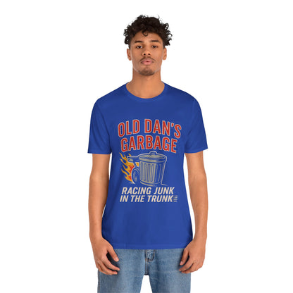 Person wearing a blue graphic T‑shirt featuring the bold red and white text “Old Dan’s Garbage” above a flaming garbage can illustration, with the tagline “Racing Junk in the Trunk” below. Funny drag racing parody shirt with retro racing flames, checkered flag background, and humor‑driven hot rod style, perfect for car enthusiasts and gearhead apparel fans.