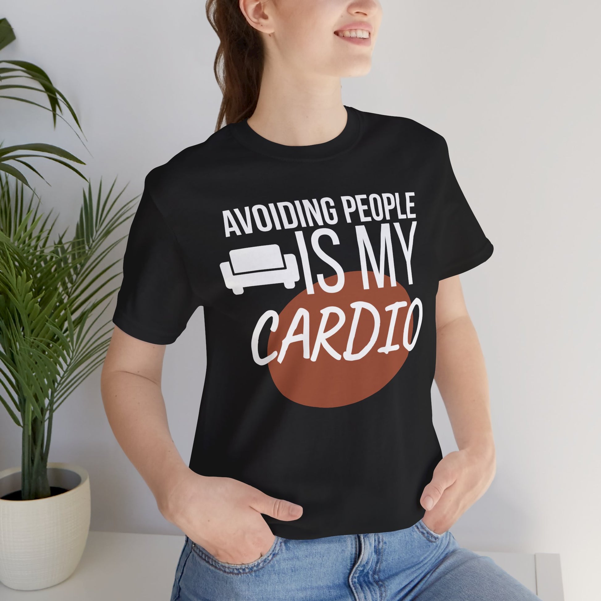 Person wearing a black t-shirt with humorous text 'Avoiding People Is My Cardio' design, standing indoors.