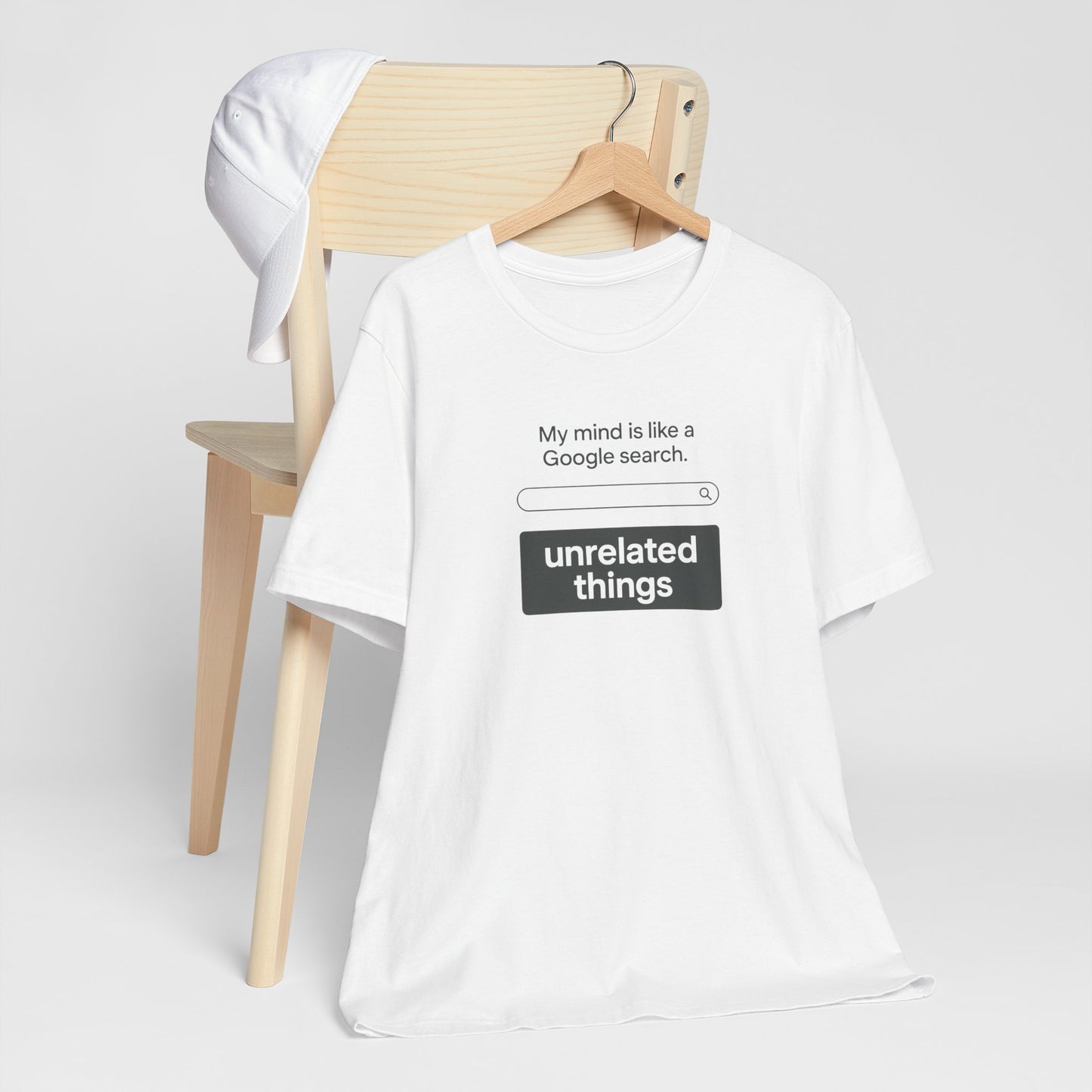 White t-shirt with humorous text design, says 'My mind is like a Google search bar' under search bar graphic, text says 'unrelated things' on a wooden chair against a white background