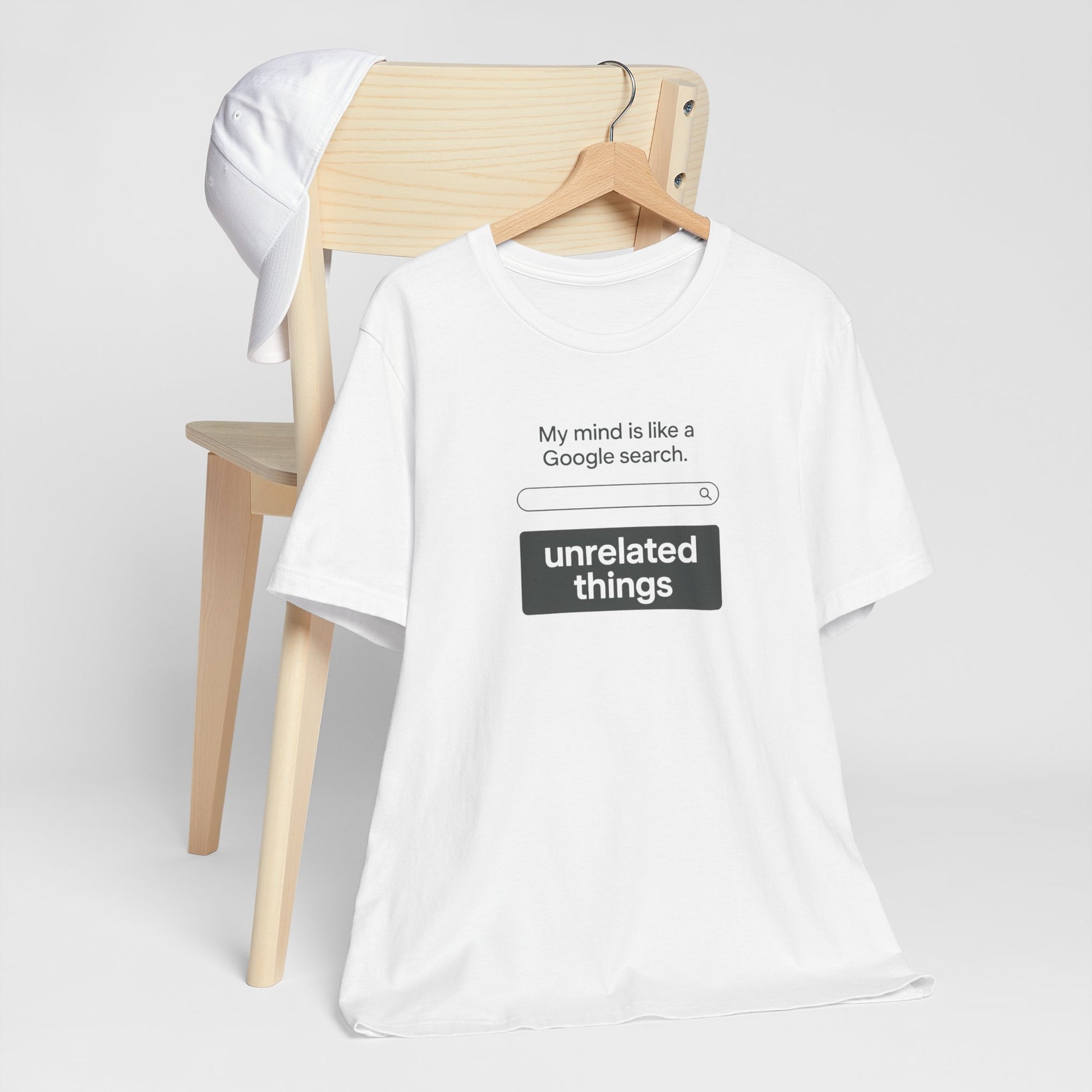 White t-shirt with humorous text design, says 'My mind is like a Google search bar' under search bar graphic, text says 'unrelated things' on a wooden chair against a white background