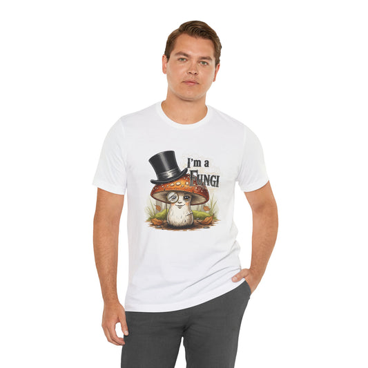 White Bella+Canvas 3001 unisex tee featuring a playful illustrated mushroom character with brown cap, white speckles, monocle, and top hat, framed by fall leaves and grass, with the pun “I’m a Fungi” in bold lettering.