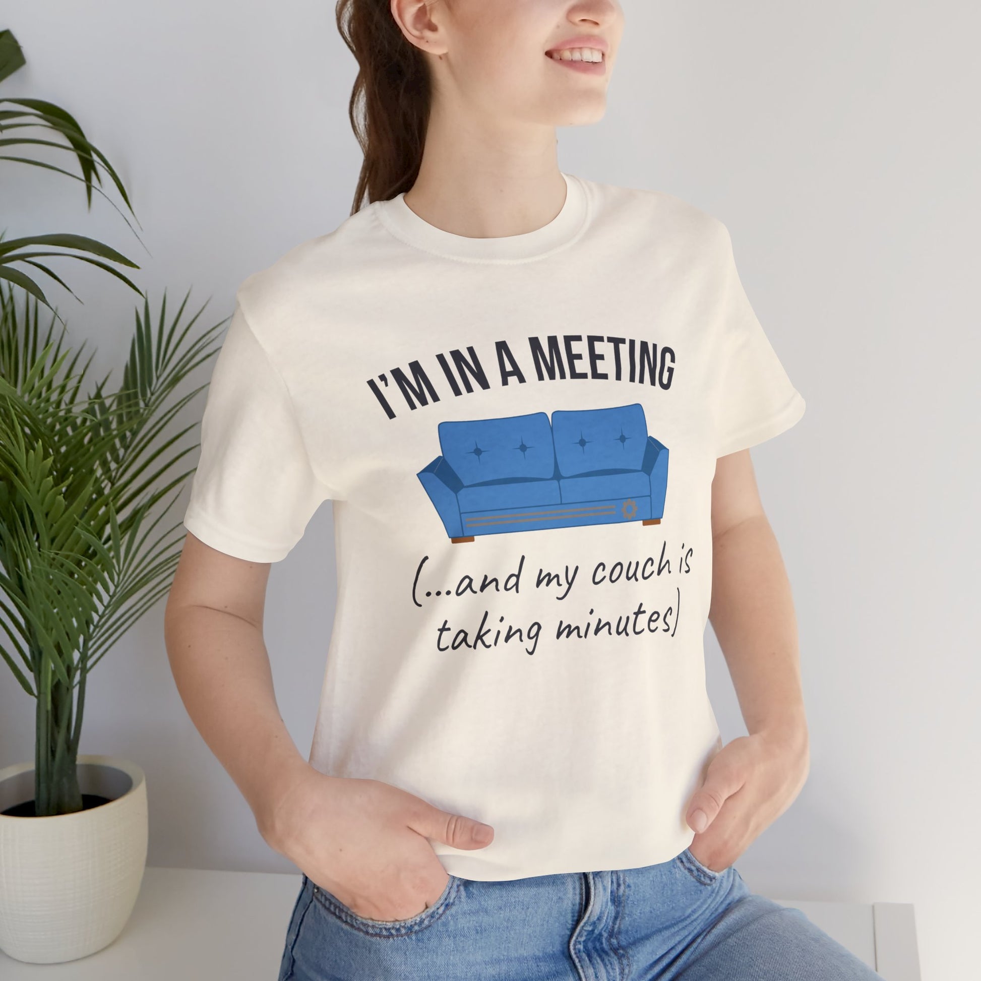 Person wearing a white t-shirt with a humorous graphic and text 'I'm in a meeting (...and my couch is taking minutes)', standing next to a plant.