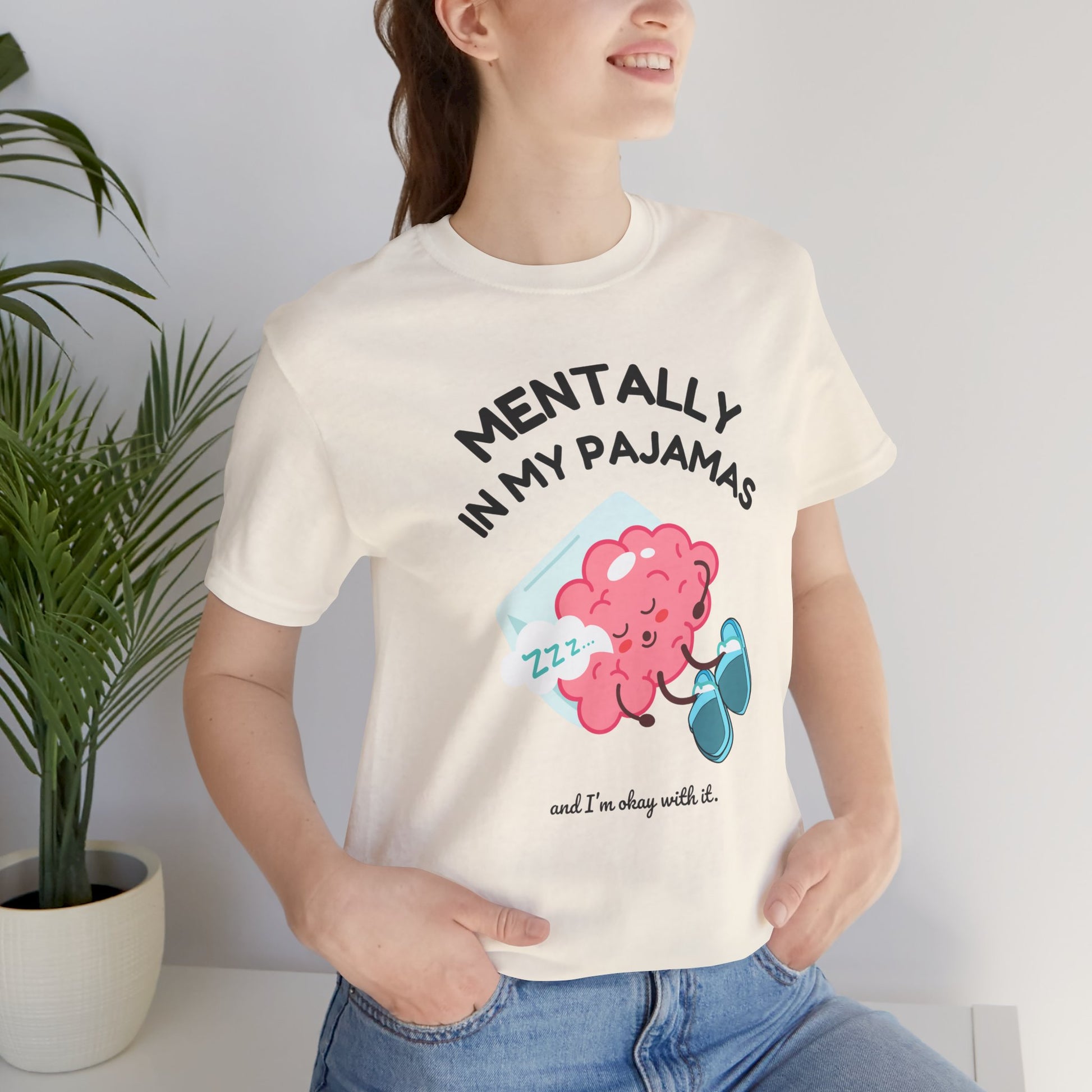 Person wearing a white t-shirt with a brain graphic and text 'Mentally in my pajamas and I'm ok with it', standing indoors next to a plant.