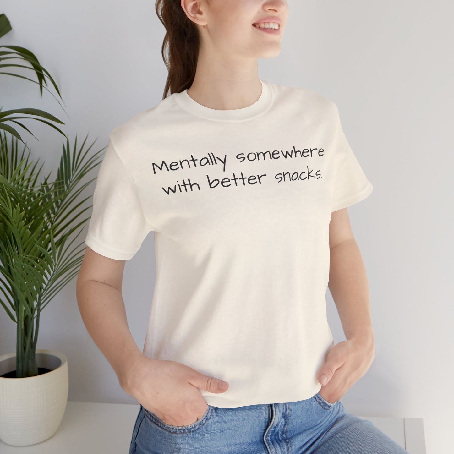Person wearing a white t-shirt with text 'Mentally somewhere with better snacks', standing next to a plant indoors.