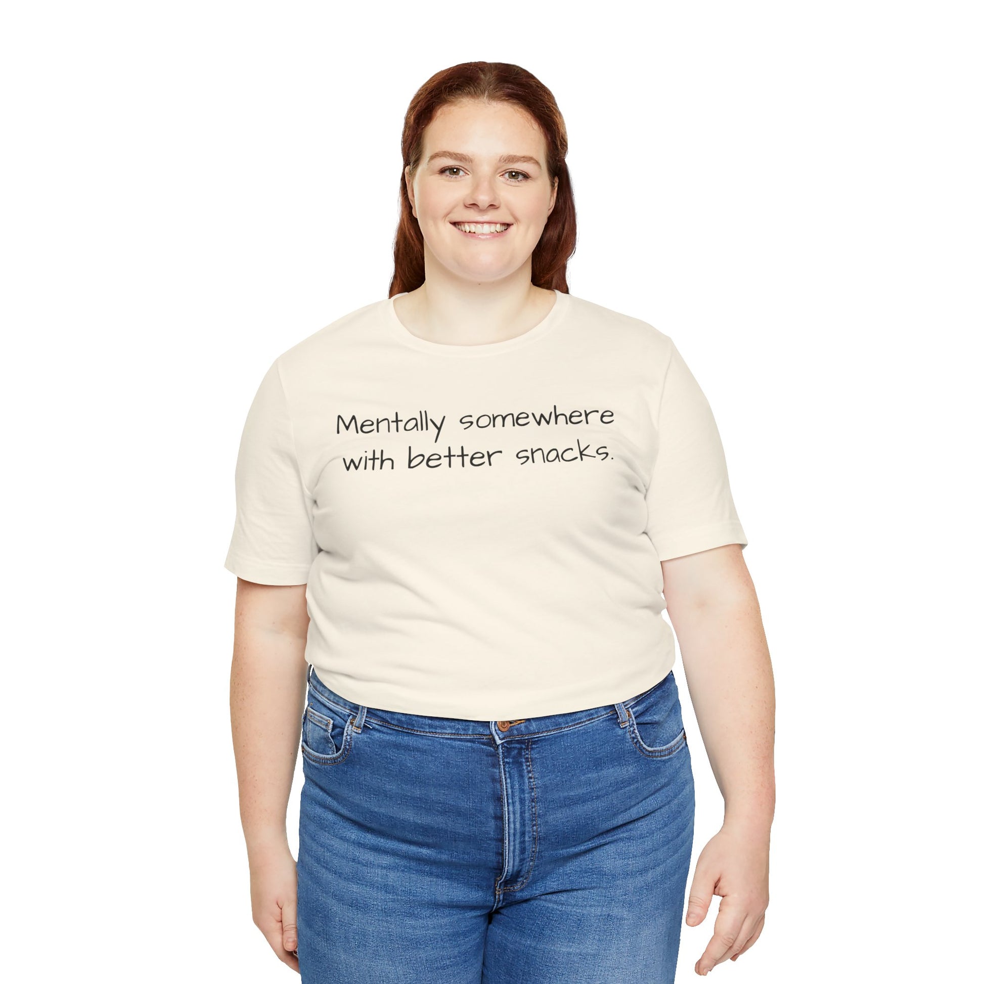 Person wearing a beige t-shirt with text 'Mentally somewhere with better snacks'   and blue jeans on a white background