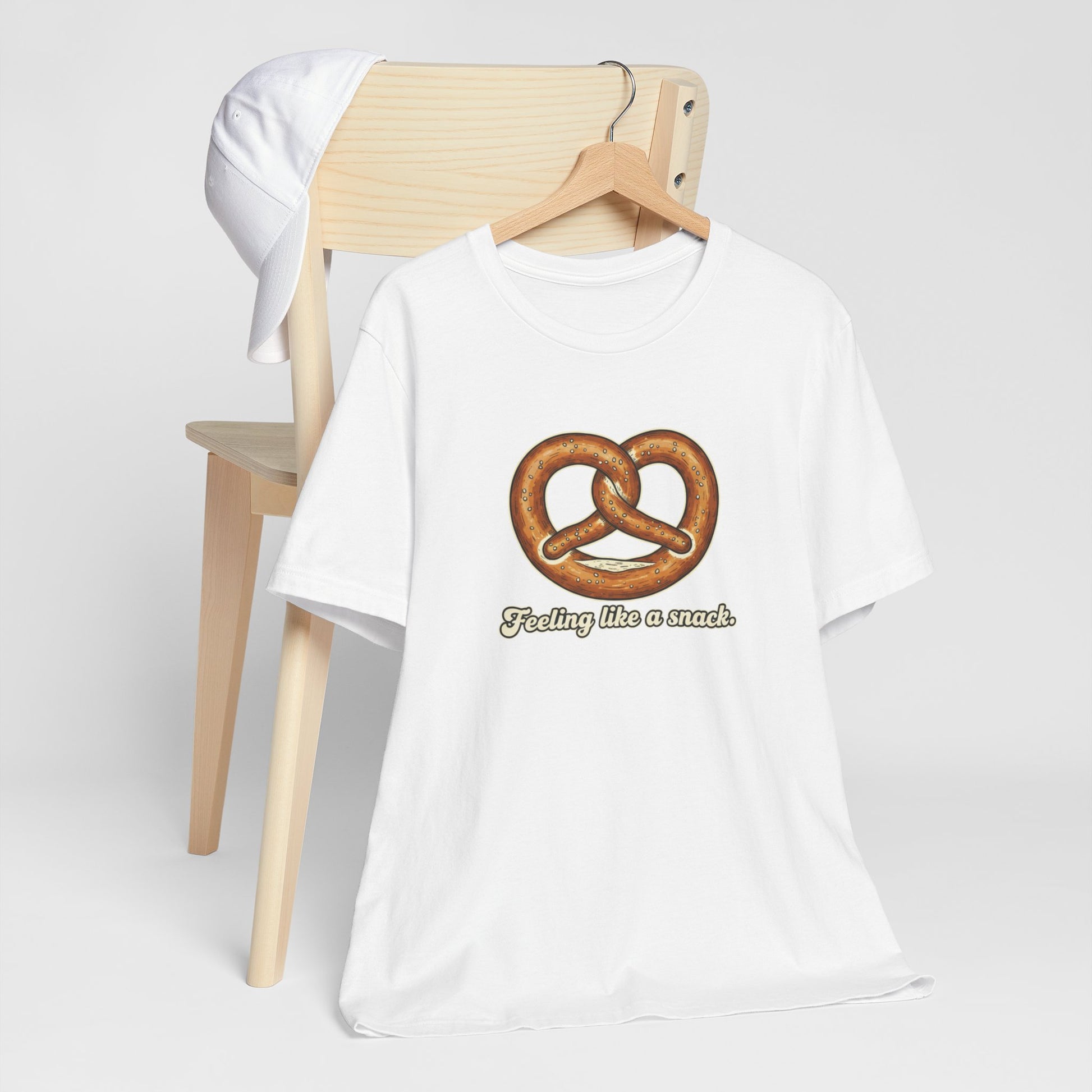 White t-shirt with pretzel graphic and text 'Feeling like a snack' on a wooden chair.