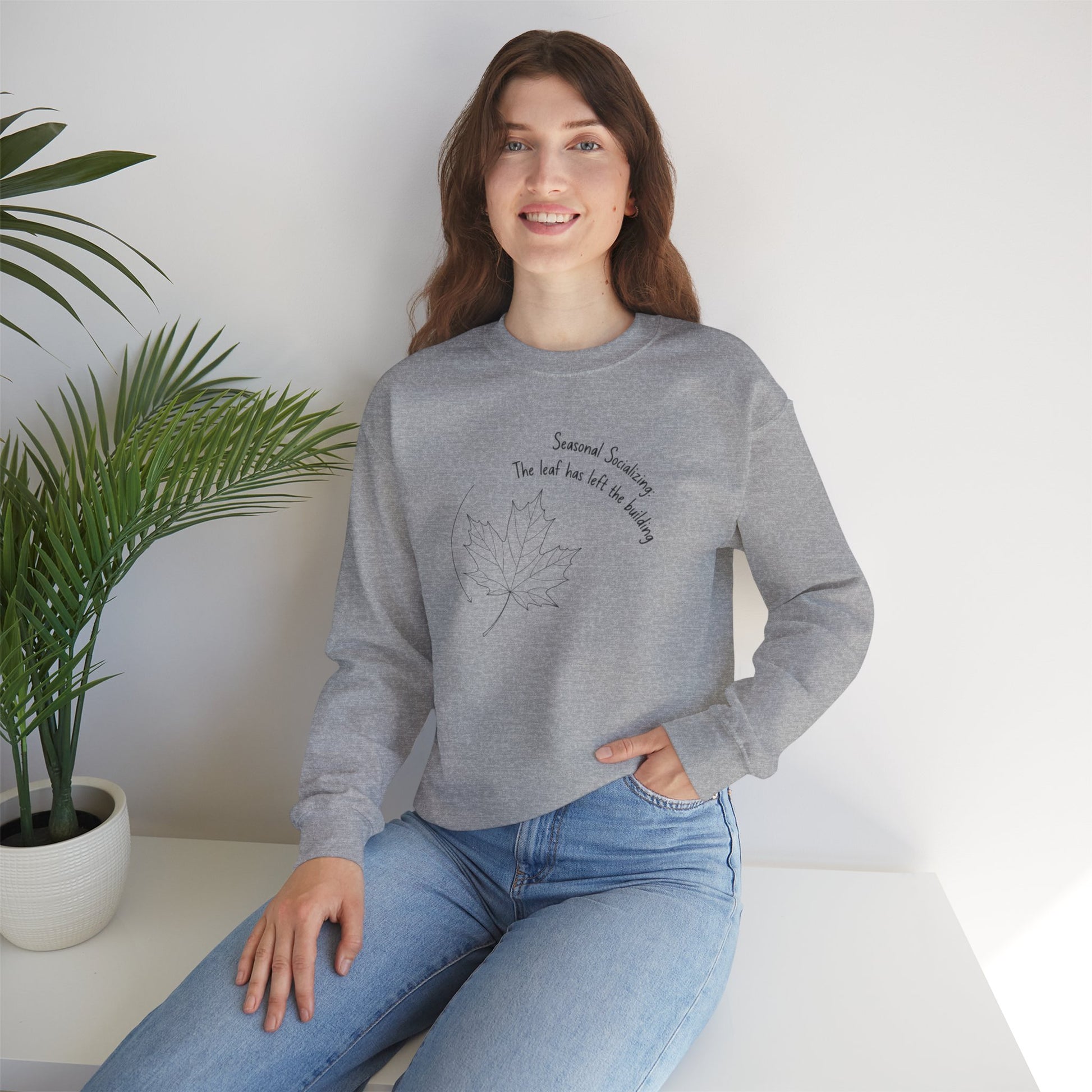 Woman wearing a gray sweatshirt with a leaf design and text 'Seasonal Socializing, The leaf has left the building', sitting on a white surface with a plant in the background.