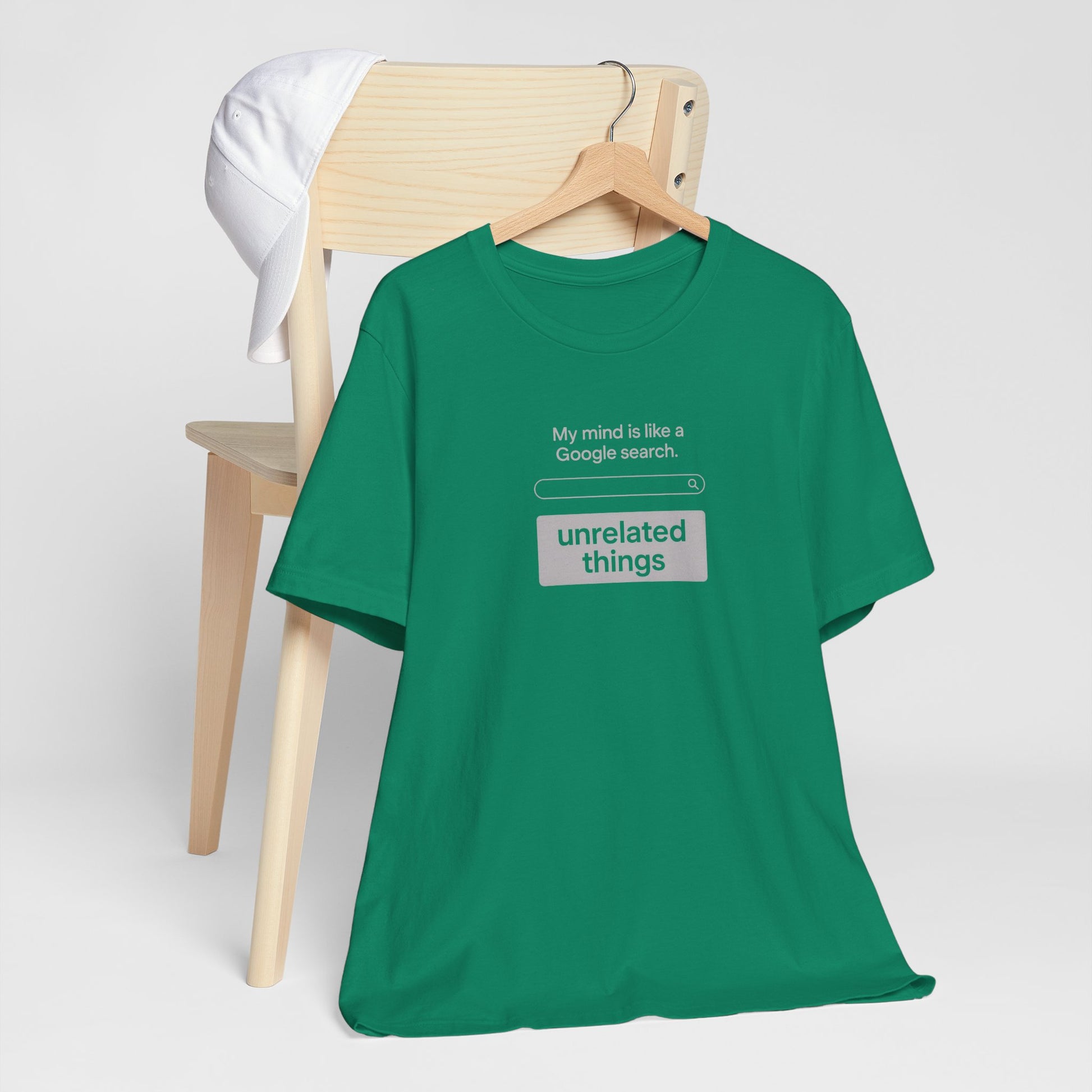 Green t-shirt with humorous text design on a wooden chair against a white background. T-shirt has a search bar graphic with text above that says, 'My mind is like a Google search' and text below that says 'unrelated things'