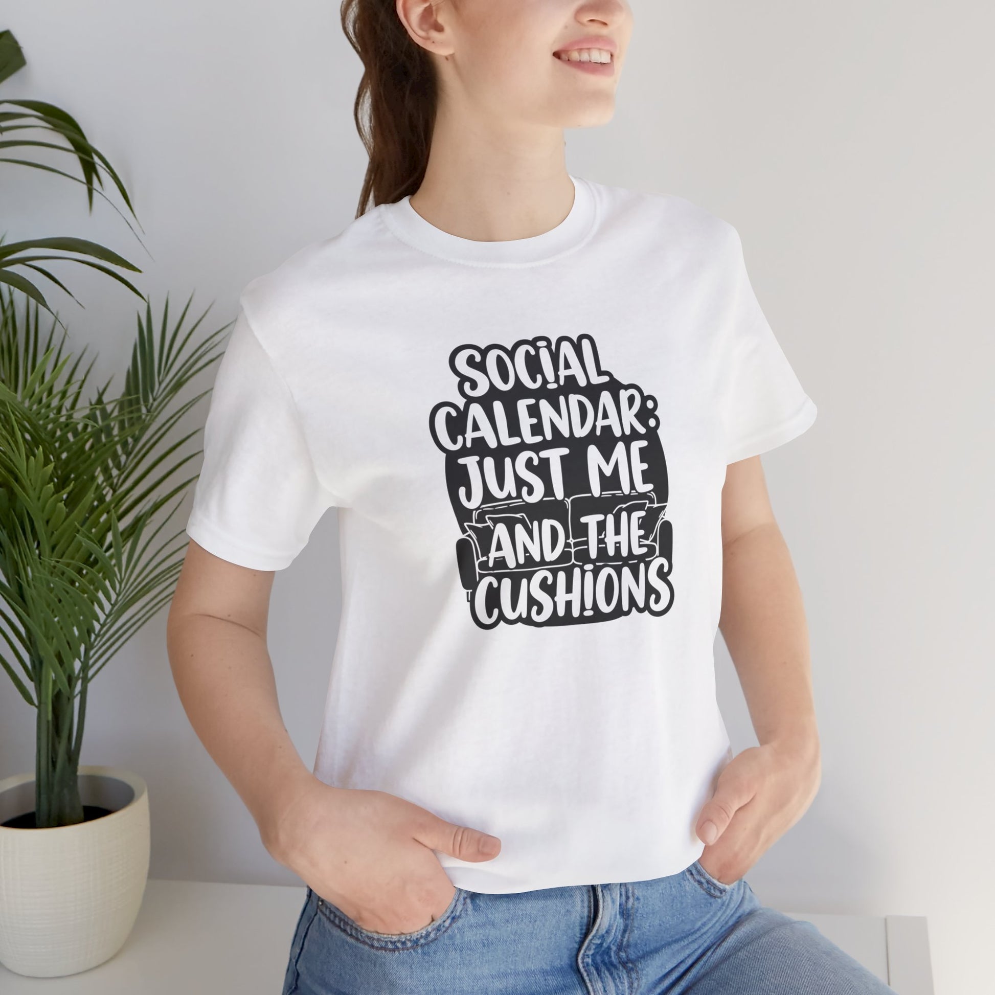 Person wearing a white t-shirt with text 'Social calendar: Just me and the cushions', standing indoors next to a plant.