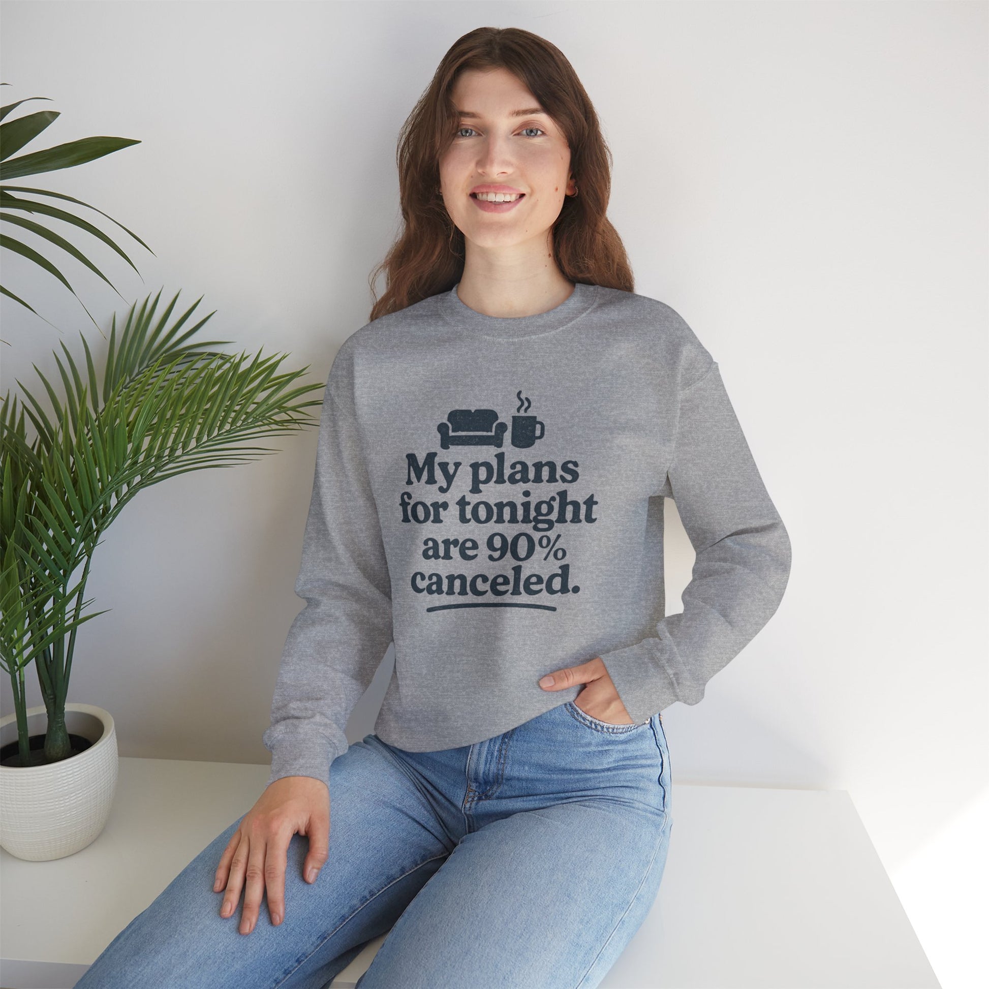 Woman wearing a gray sweatshirt with text 'My plans for tonight are 90% canceled' sitting on a white surface with a plant in the background
