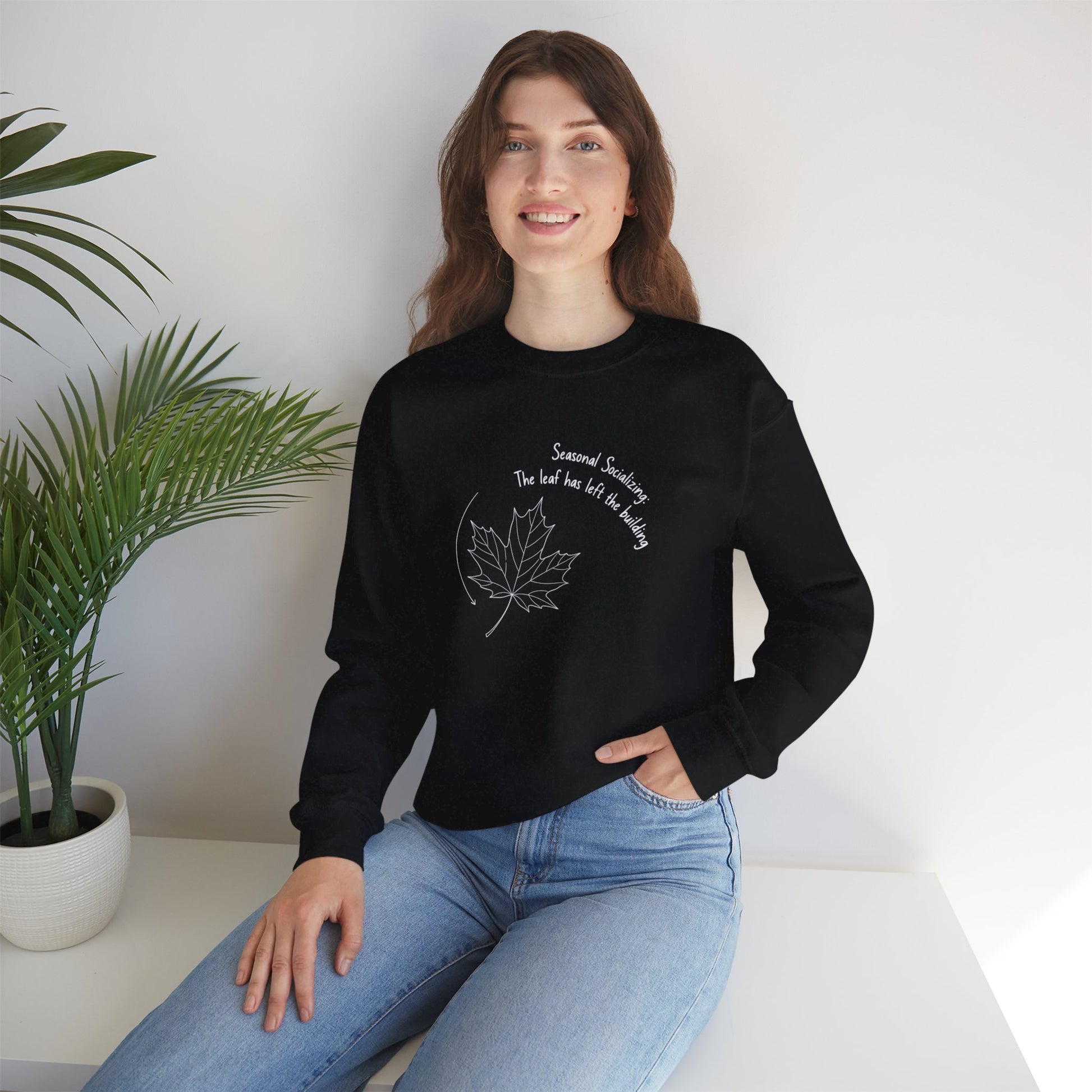 Person wearing a black sweatshirt with a leaf design and text 'Seasonal Socializing, The leaf has left the building', sitting on a white surface with a plant in the background.