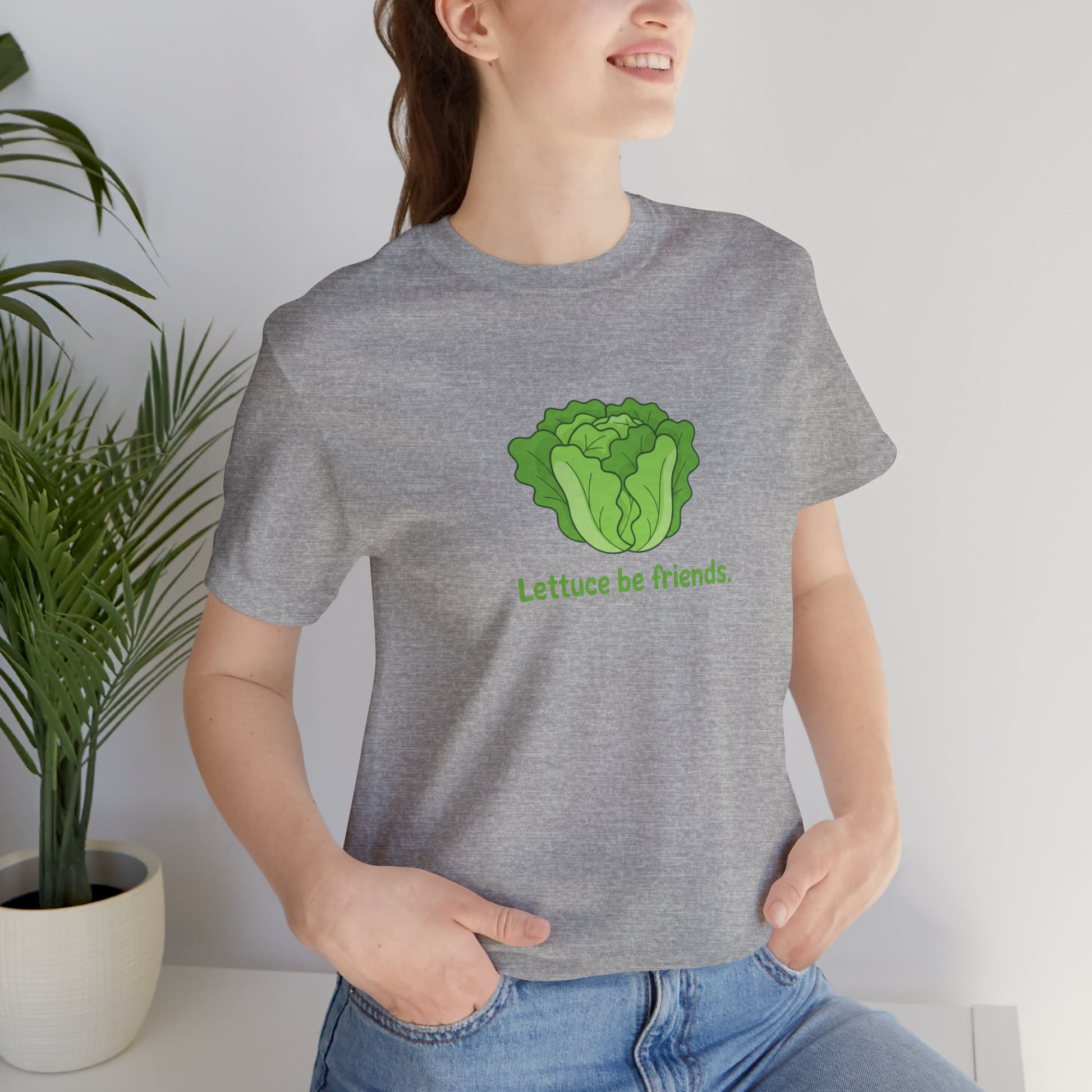 Person wearing a gray t-shirt with a green lettuce graphic and text 'Lettuce be friends', standing indoors next to a plant.