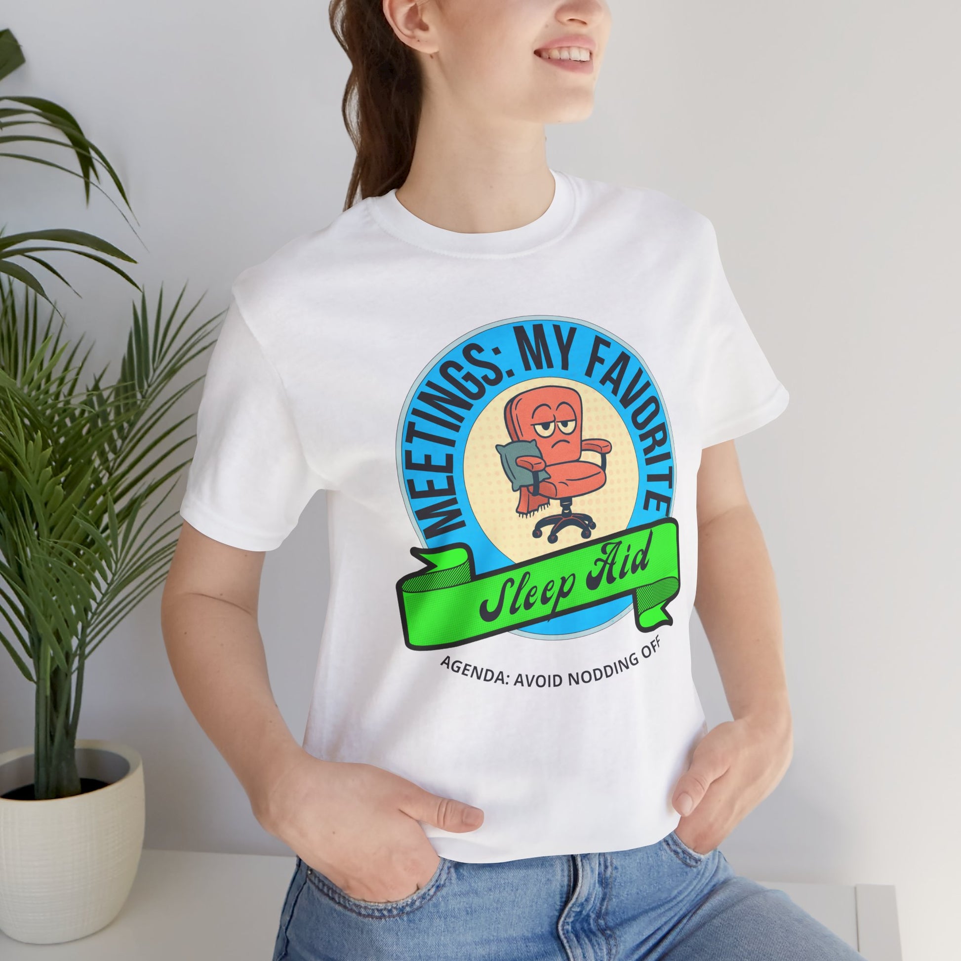 Person wearing a white t-shirt with a colorful graphic design and text 'Meetings: My favorite sleep aid. Agenda: Avoid nodding off', standing indoors next to a plant.