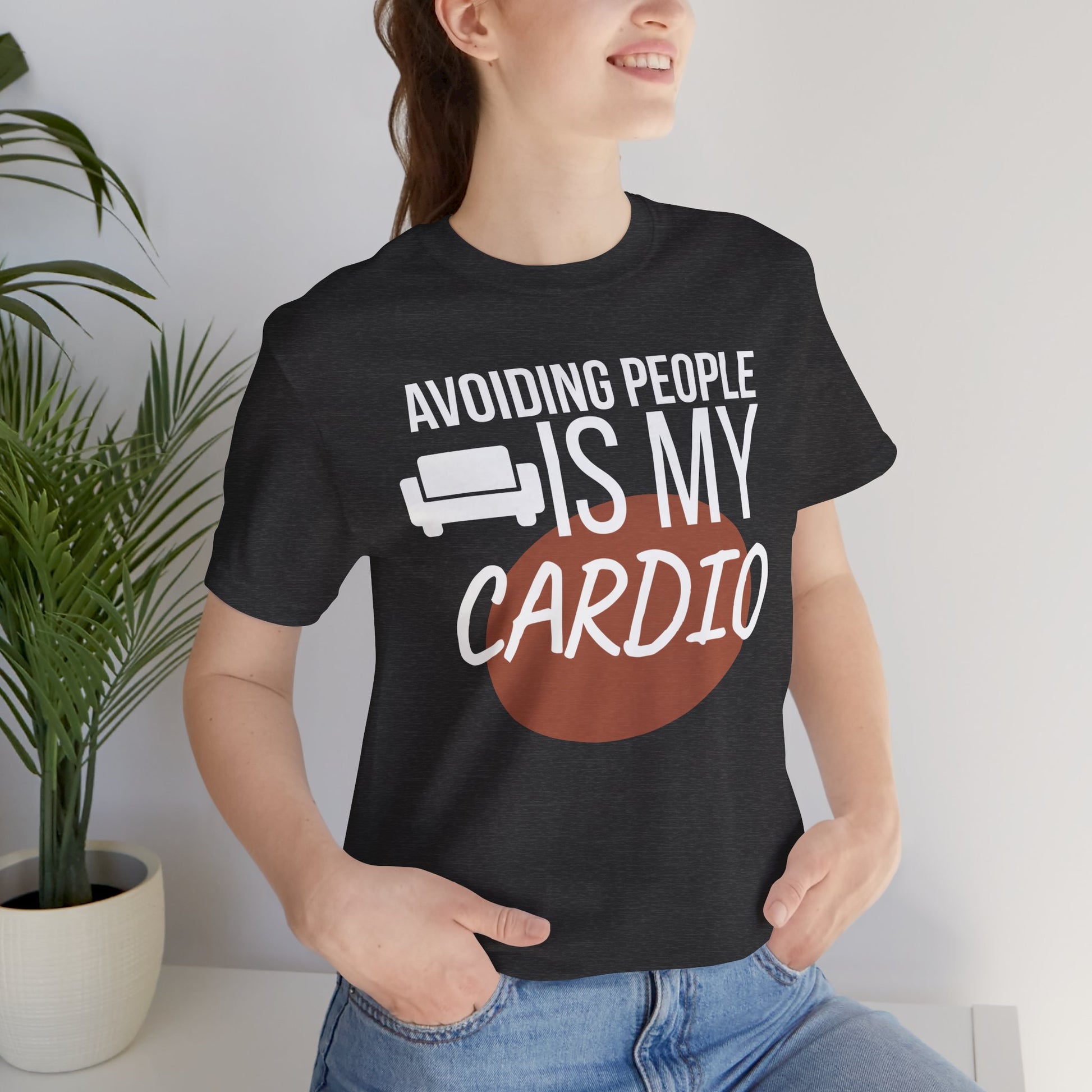 Person wearing a black t-shirt with humorous text 'Avoiding People Is My Cardio' design, standing indoors.