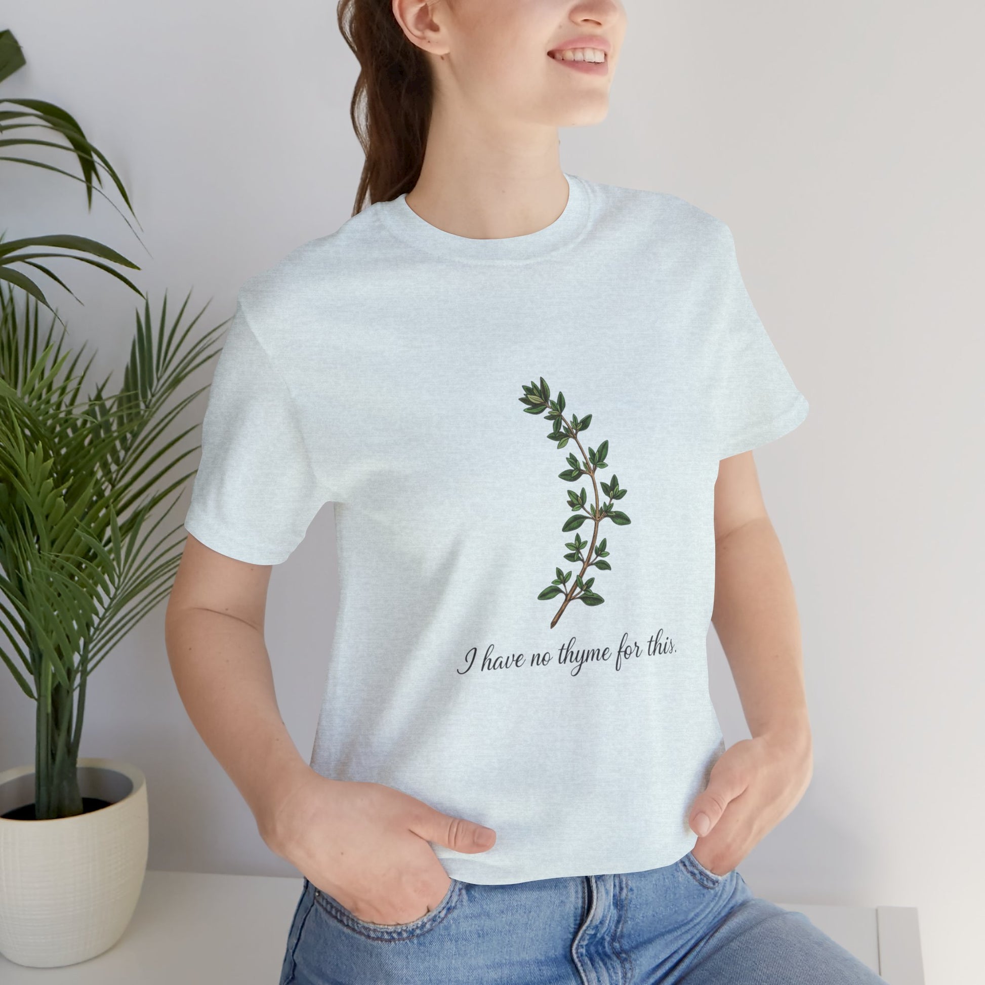 Person wearing a white t-shirt with a plant design and text 'I have no thyme for this', standing indoors next to a plant.