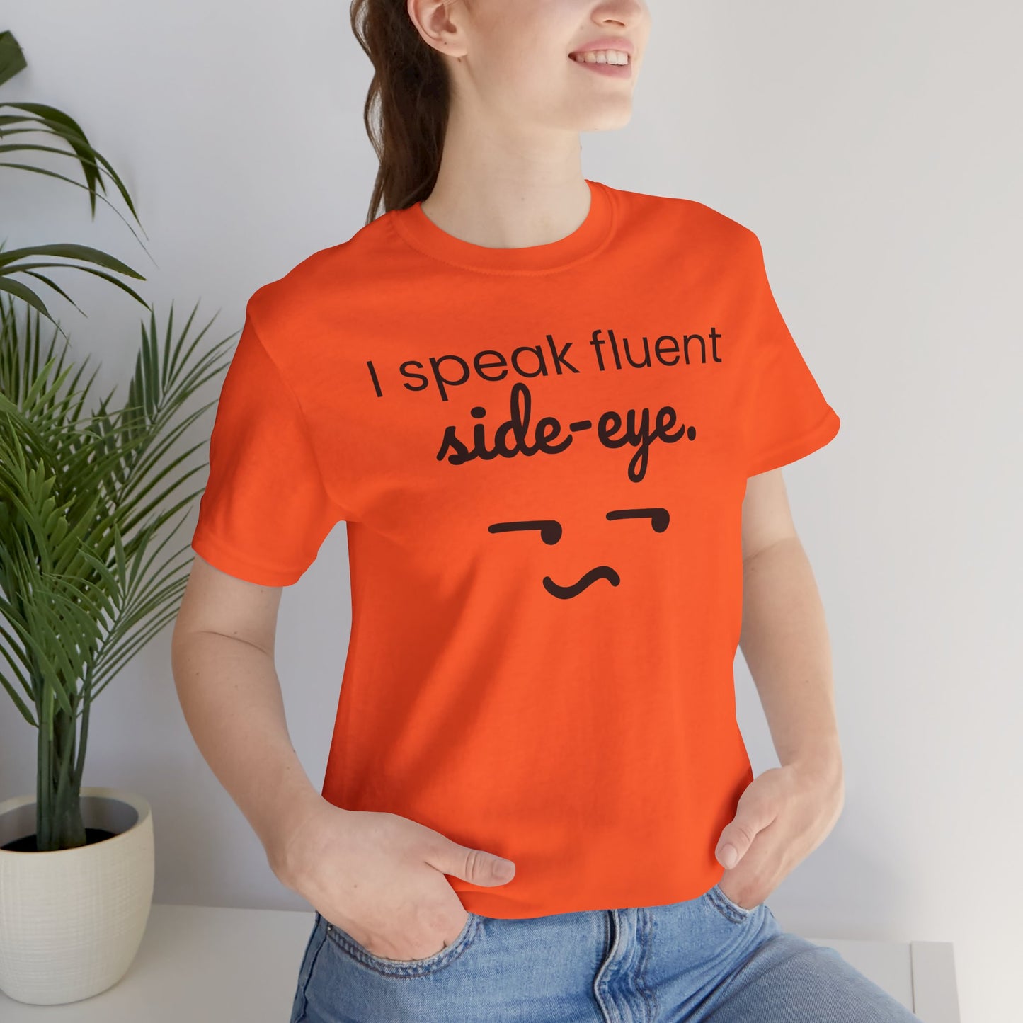 Person wearing an orange t-shirt with text 'I speak fluent side eye' and a face graphic, standing against a white wall with a plant.