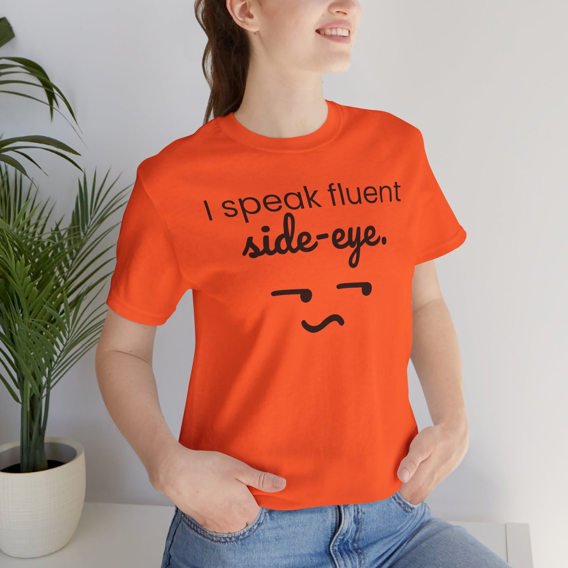 Person wearing an orange t-shirt with text 'I speak fluent side eye' and a face graphic, standing against a white wall with a plant.