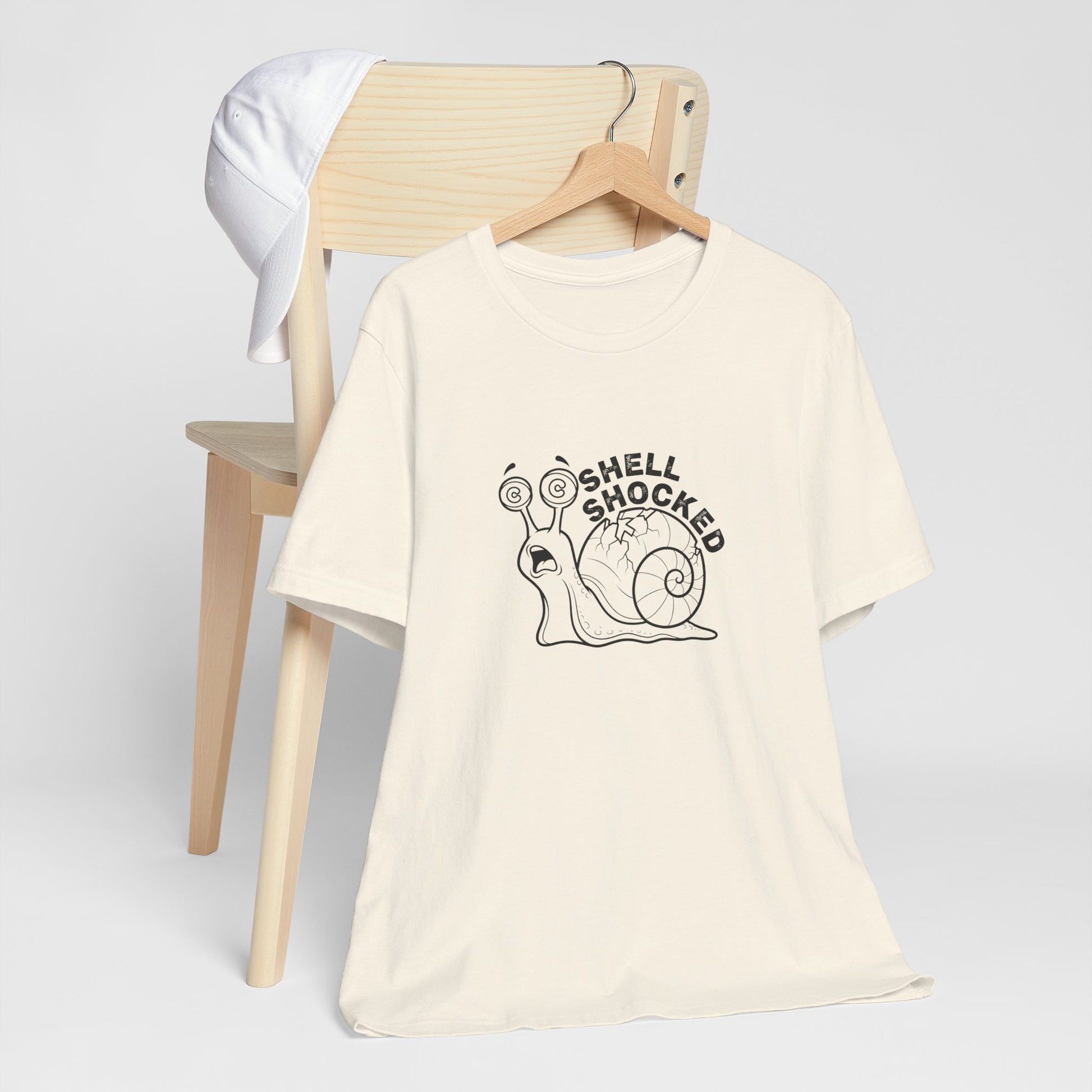 Beige t-shirt with 'Shell Shocked' graphic on a wooden chair against a white background
