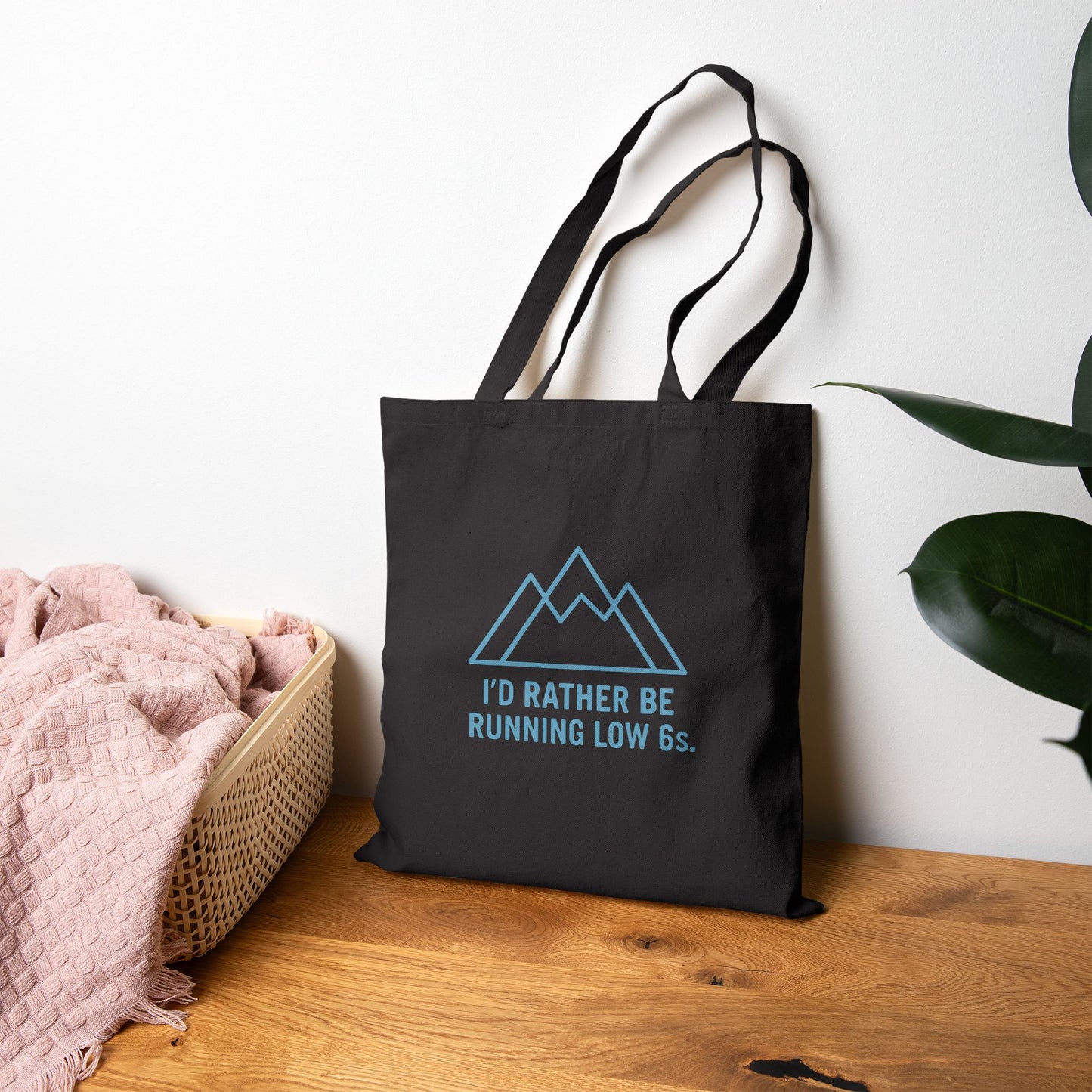 Black tote bag with blue graphic and text 'I'd rather be running low 6s' on a wooden surface with a white wall and plant in the background