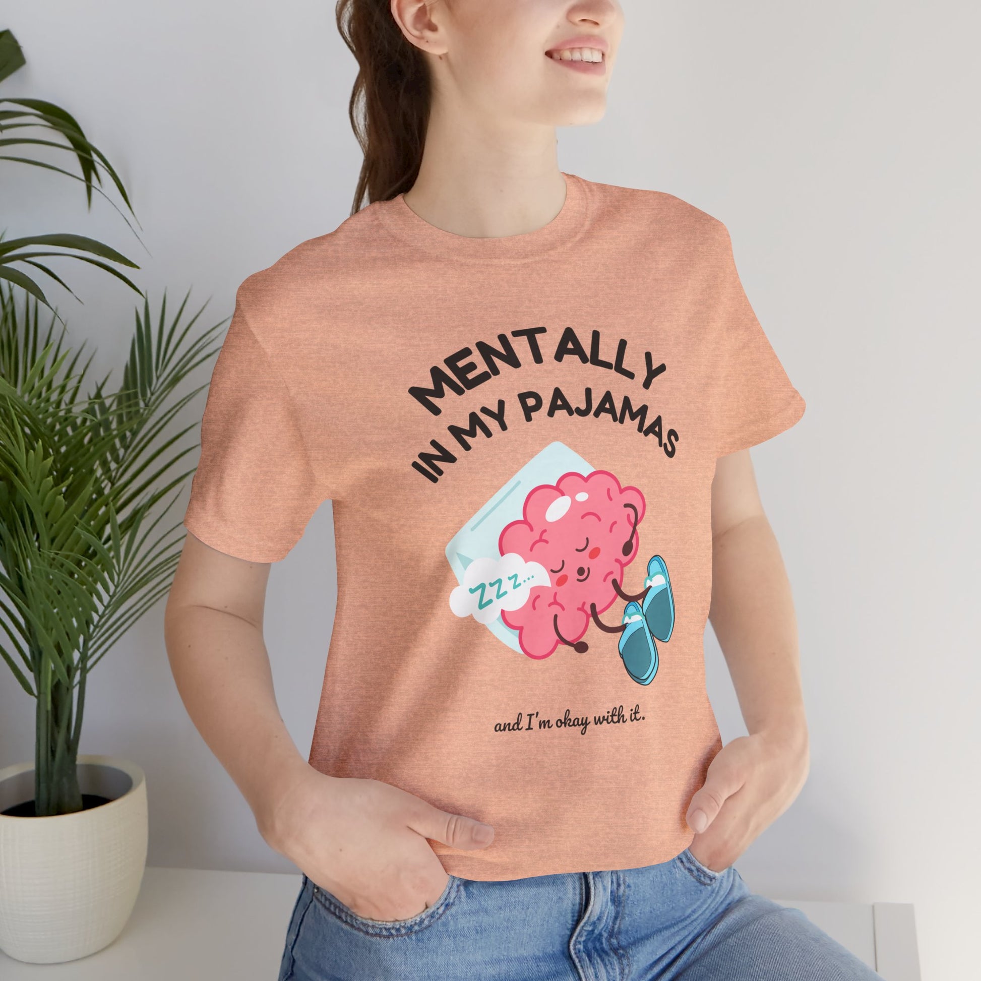 Person wearing a peach t-shirt with a brain graphic and text 'Mentally in my pajamas and I'm ok with it', standing indoors next to a plant.