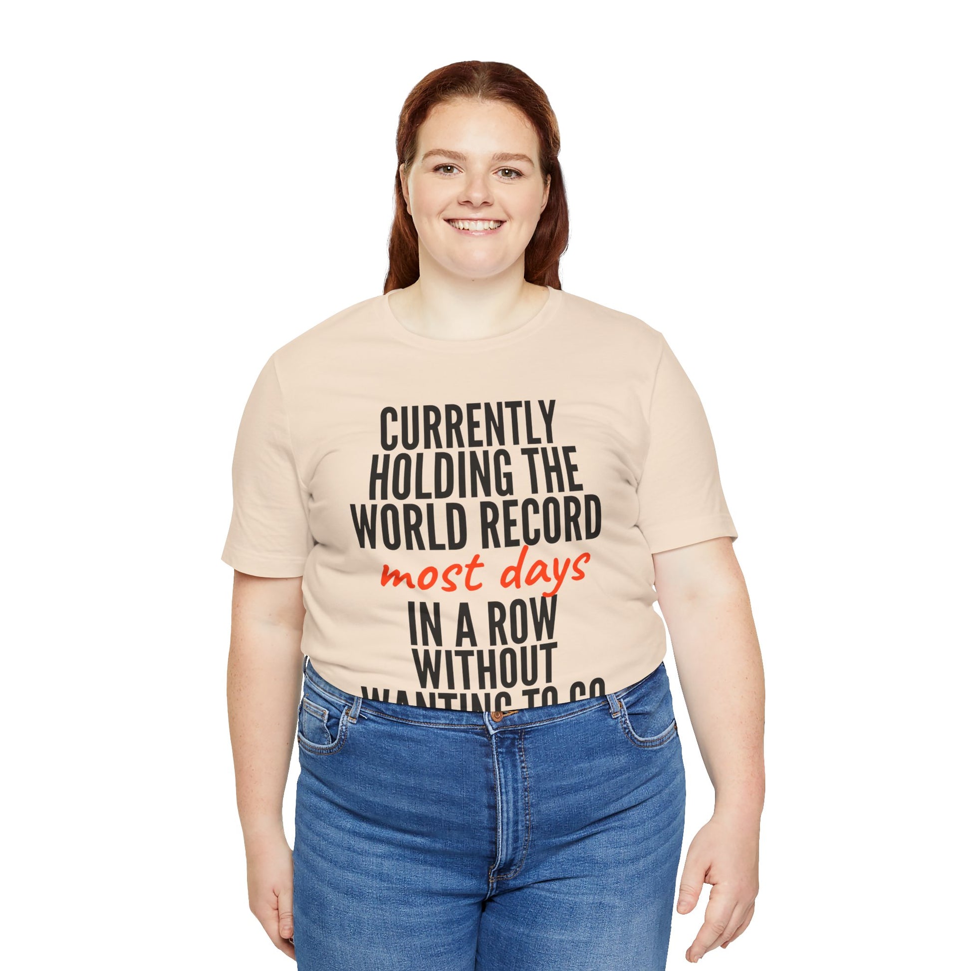 Person wearing a beige t-shirt with humorous text 'Currently holding the world record most days in a row without wanting to go' design on a white background