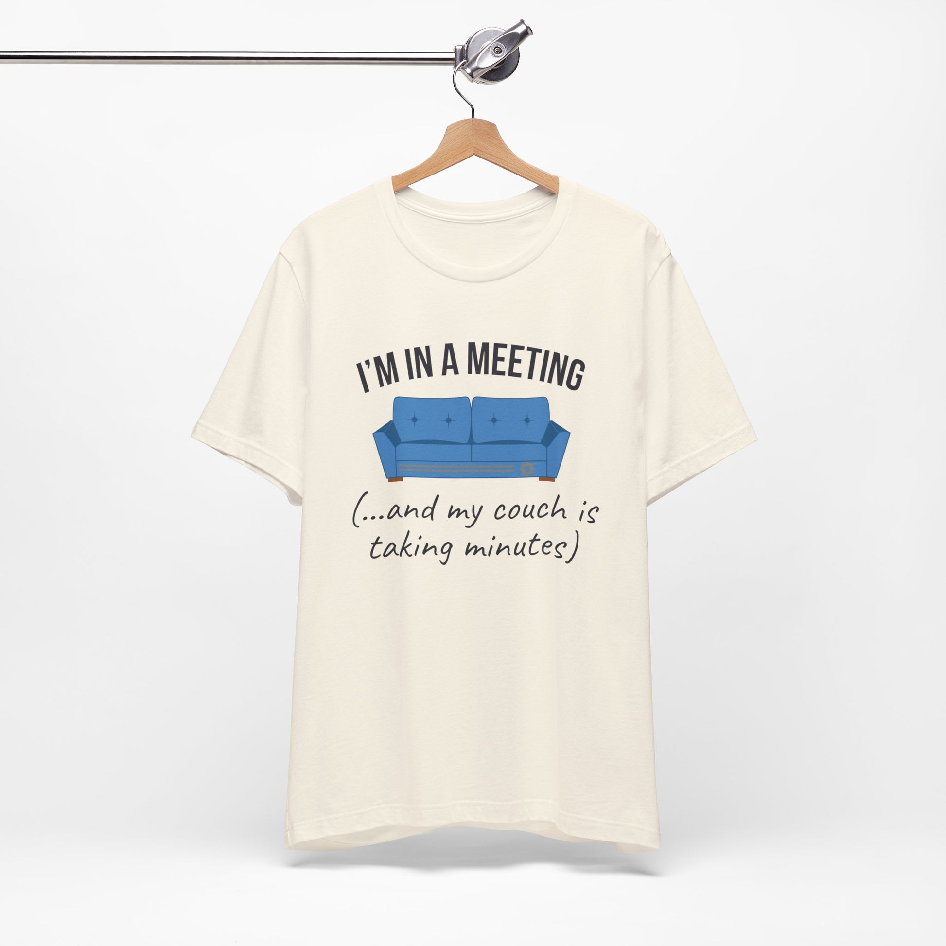 Natural t-shirt with humorous graphic and text 'I'm in a meeting (...and my couch is taking minutes)' on a hanger against a white background
