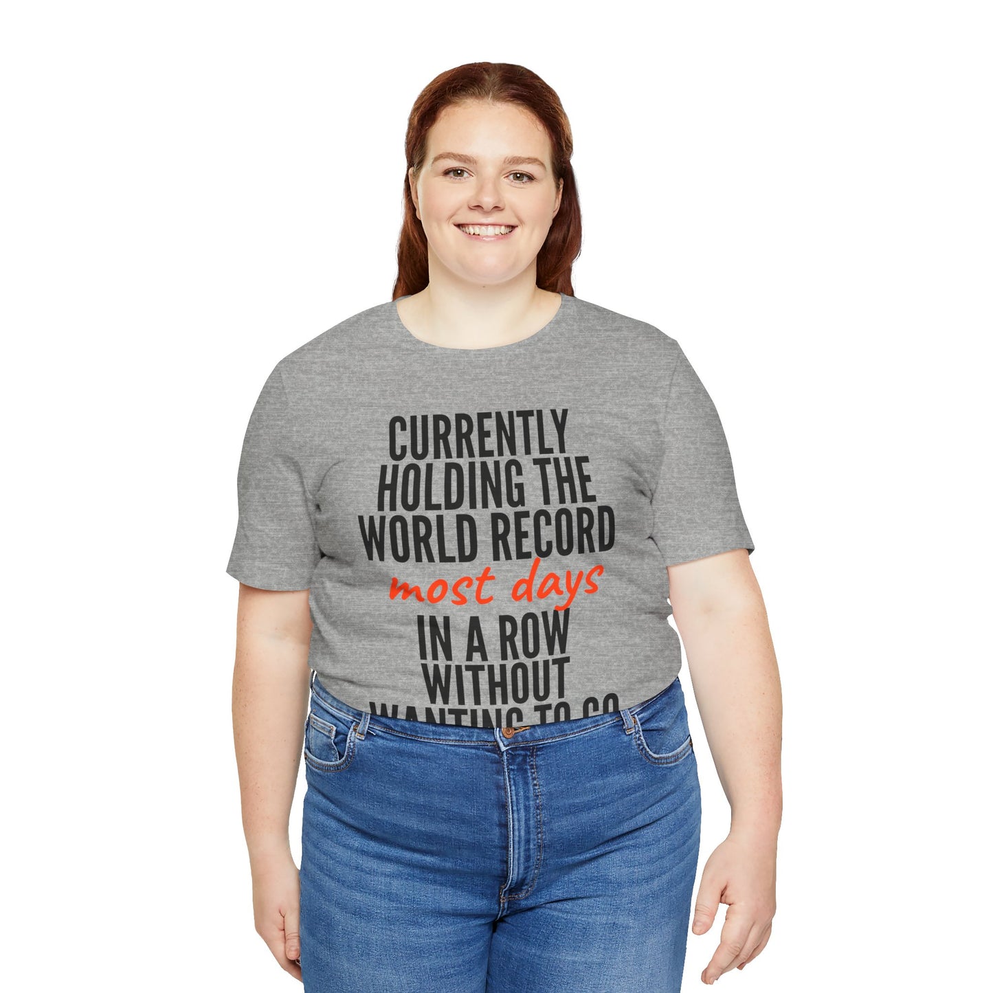 Person wearing a gray t-shirt with humorous text 'Currently holding the world record most days in a row without wanting to go' design on a white background