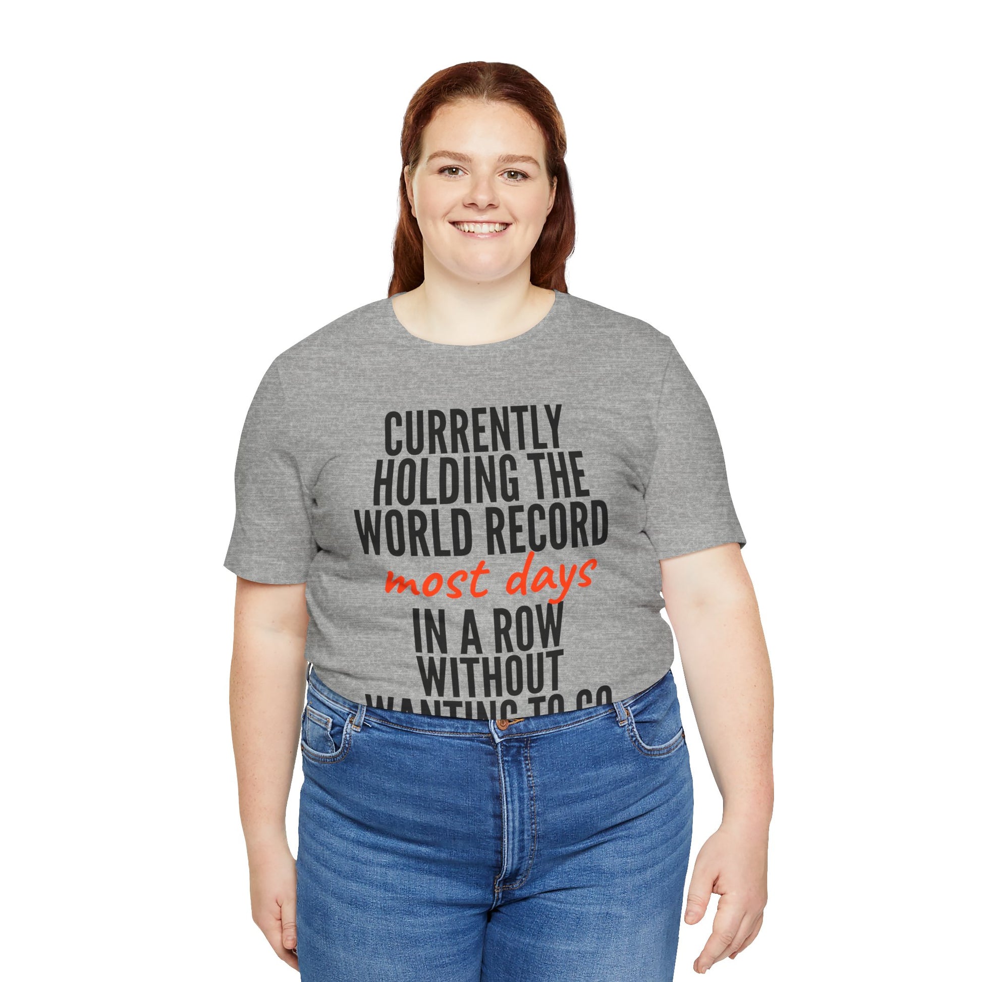 Person wearing a gray t-shirt with humorous text 'Currently holding the world record most days in a row without wanting to go' design on a white background