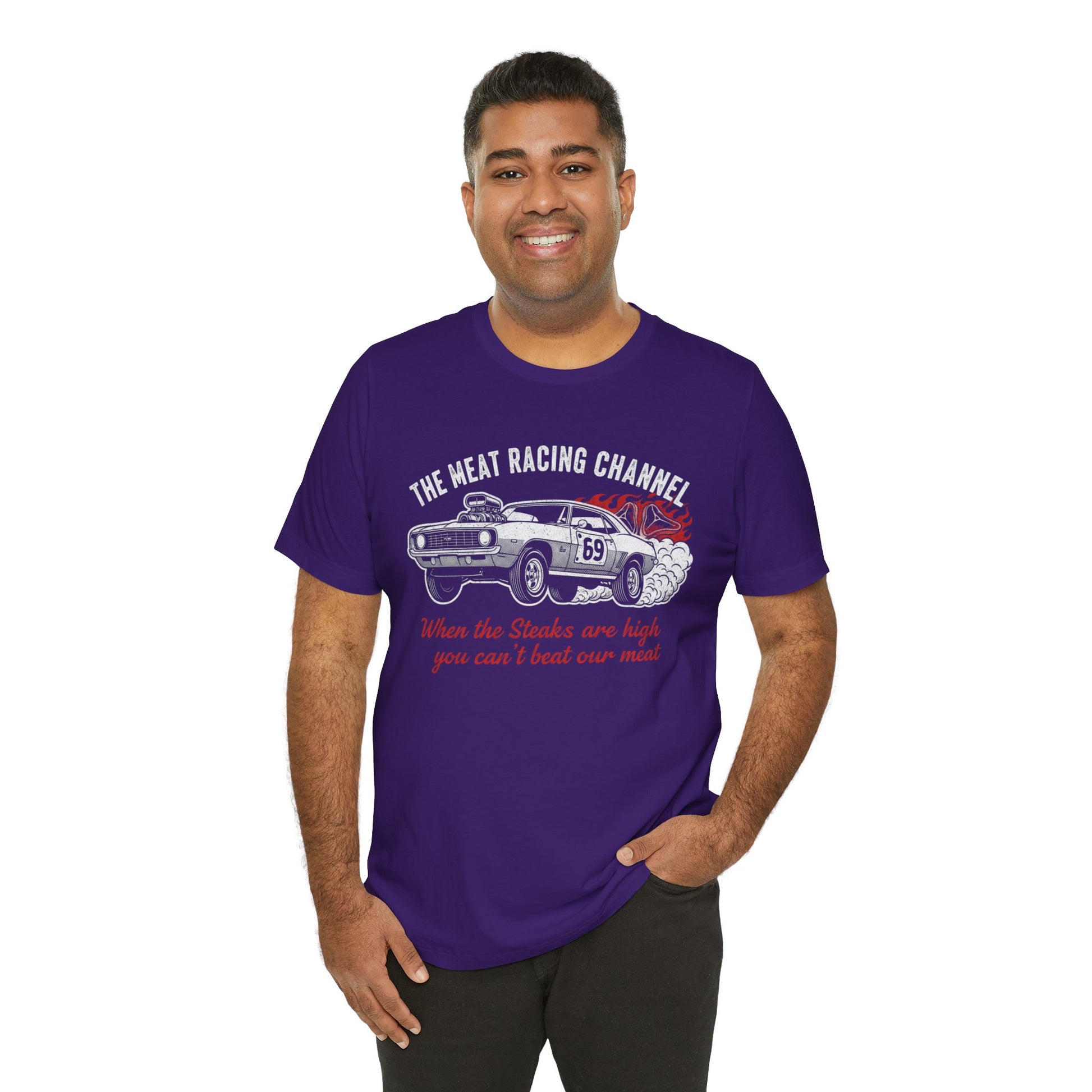 Person wearing a purple Bella+Canvas 3001 T‑shirt featuring a bold drag racing graphic. The design shows a vintage muscle car with the number 69, white smoke and red flames, the distressed white title “The Meat Racing Channel” above, and the red script tagline “When the Steaks are high you can’t beat our meat” below. Funny drag racing parody shirt with retro hot rod style, perfect for car enthusiasts, gearheads, and humor‑driven racing apparel fans.