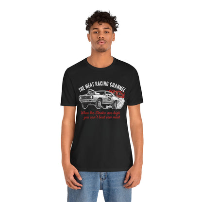 Person wearing a black Bella+Canvas 3001 T‑shirt featuring a bold drag racing graphic. The design shows a vintage muscle car with the number 69, white smoke and red flames, the distressed white title “The Meat Racing Channel” above, and the red script tagline “When the Steaks are high you can’t beat our meat” below. Funny drag racing parody shirt with retro hot rod style, perfect for car enthusiasts, gearheads, and humor‑driven racing apparel fans.