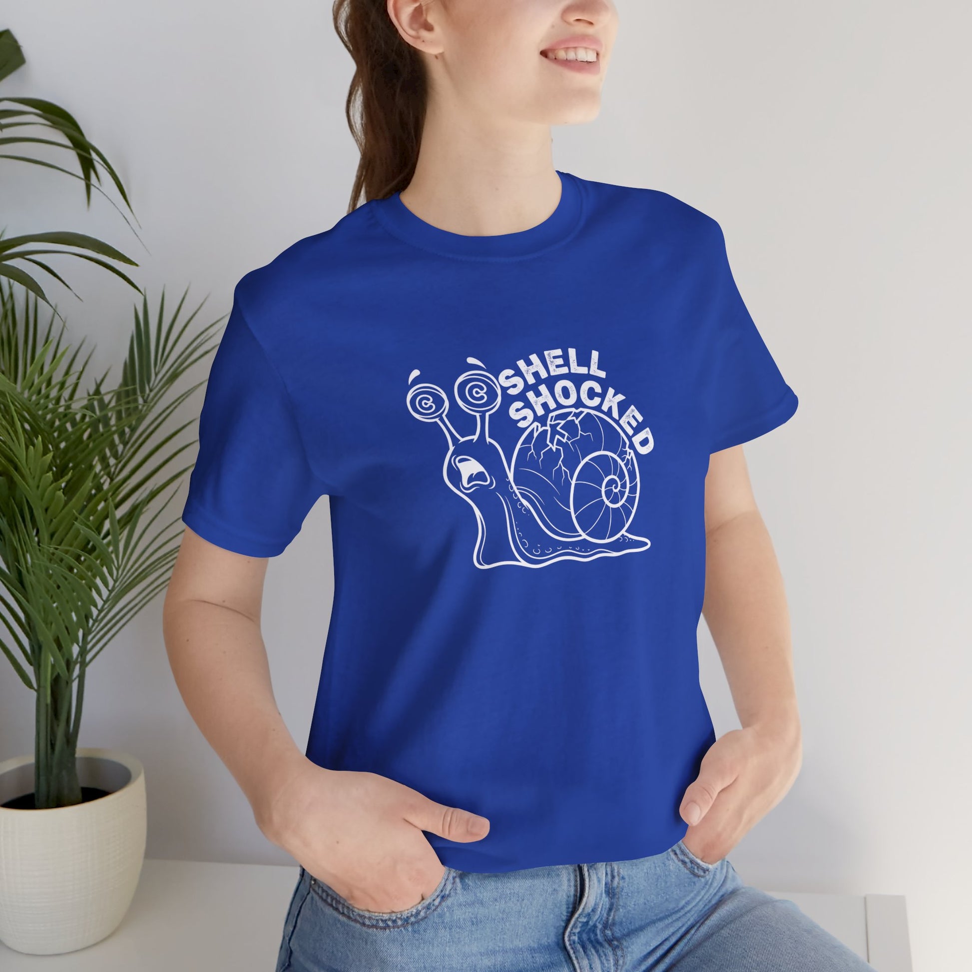 Person wearing a blue t-shirt with a snail graphic and 'Shell Shocked' text, standing indoors.