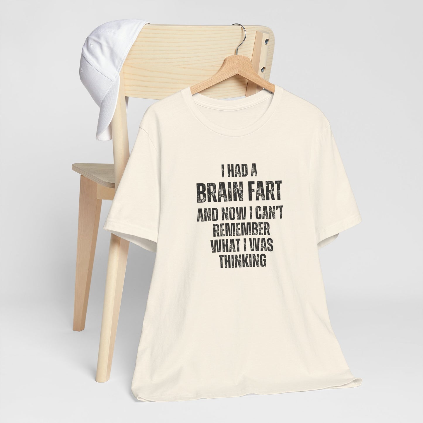 Beige t-shirt with humorous text 'I had a brain fart and now I can't remember what I was thinking' on a wooden chair against a white background