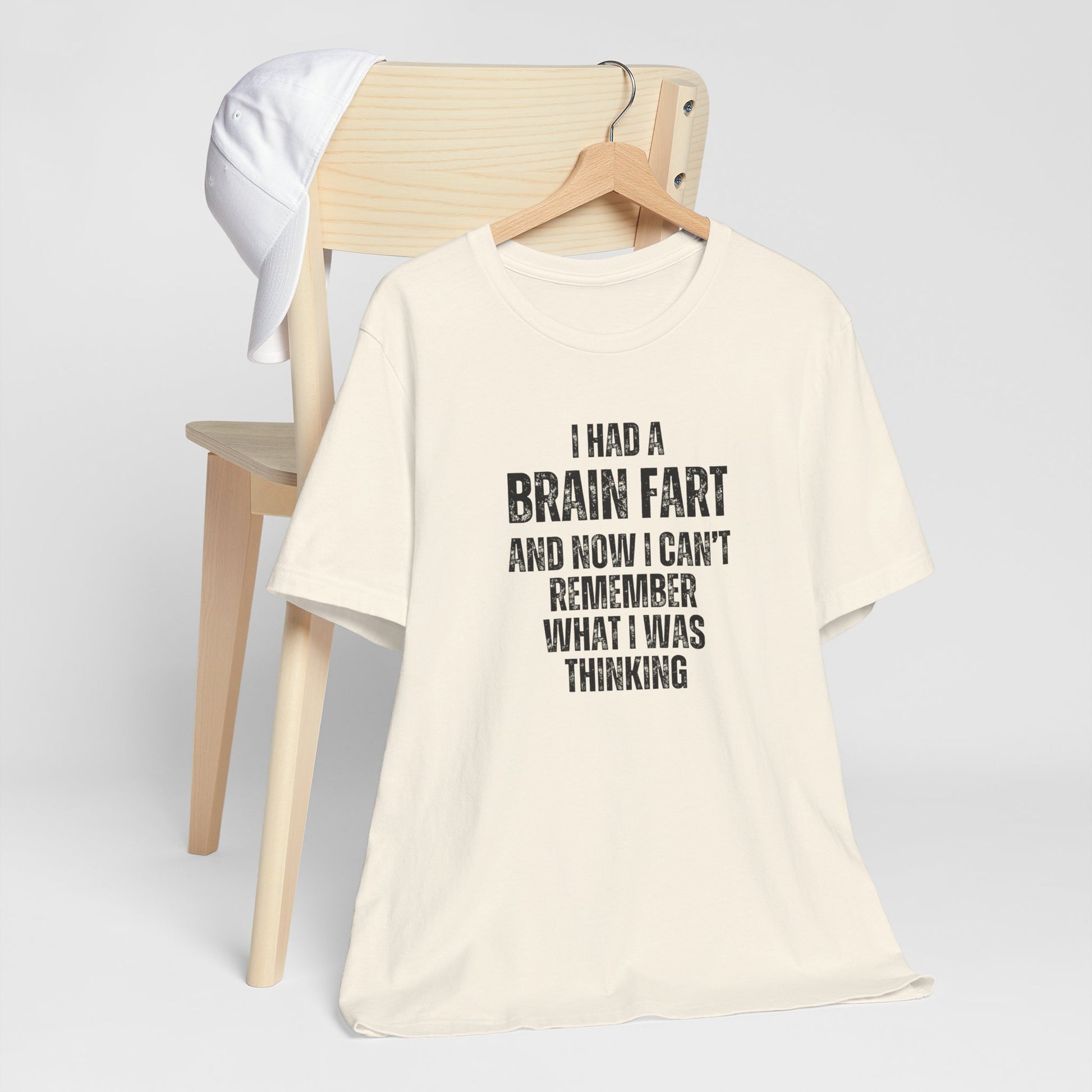 Beige t-shirt with humorous text 'I had a brain fart and now I can't remember what I was thinking' on a wooden chair against a white background