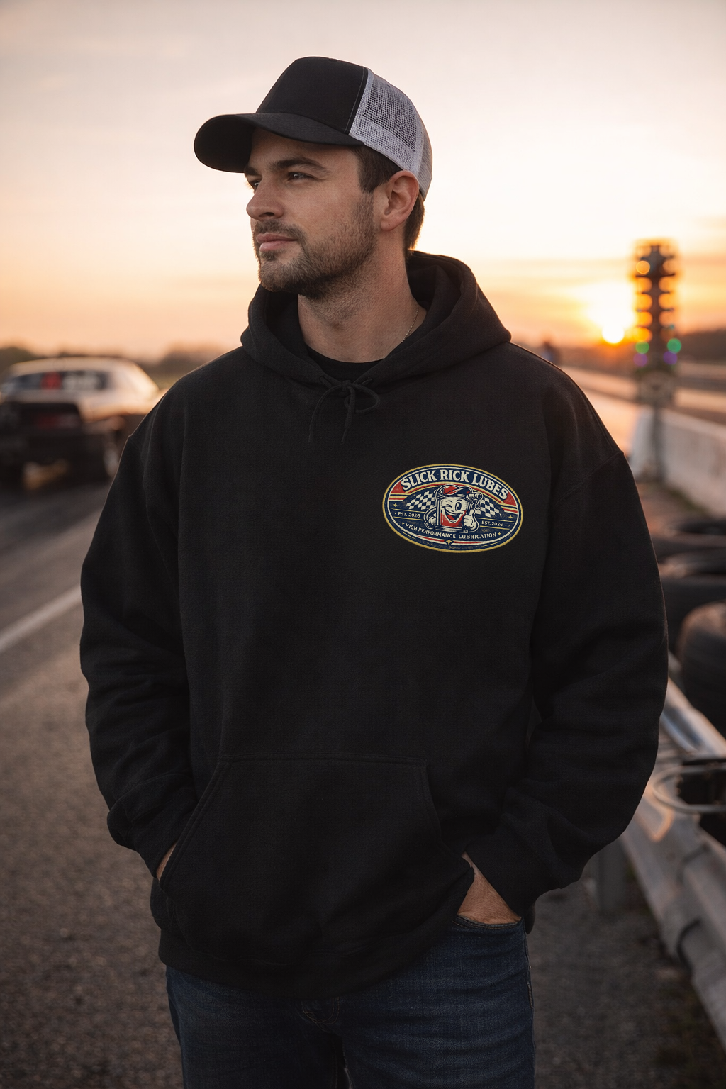 Man wearing a black hoodie with a 'Slick Rick Luges' logo, standing on a road during sunset.