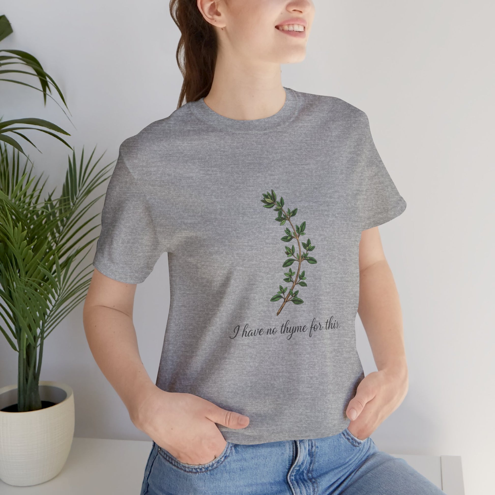 Person wearing a gray t-shirt with a plant design and text 'I have no thyme for this', standing next to a potted plant.