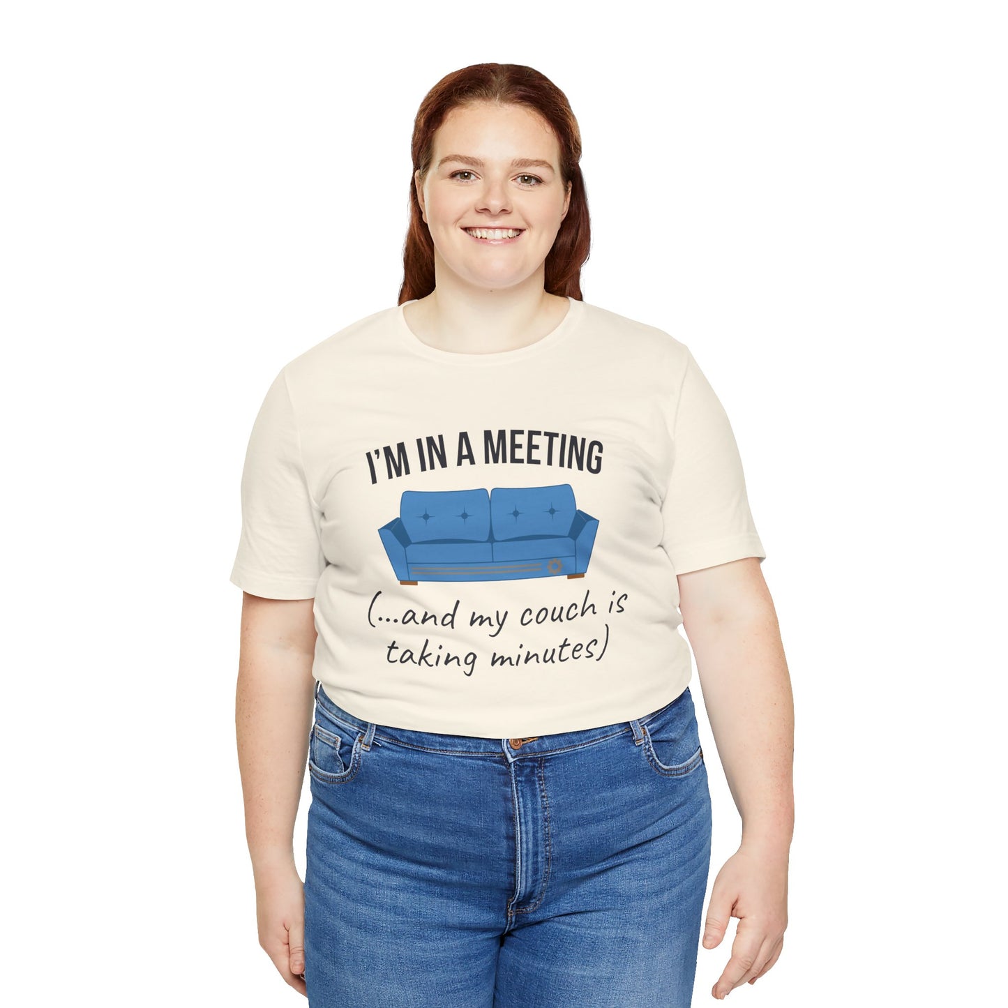 Person wearing a cream t-shirt with a graphic and text 'I'm in a meeting (...and my couch is taking minutes)', standing against a white background