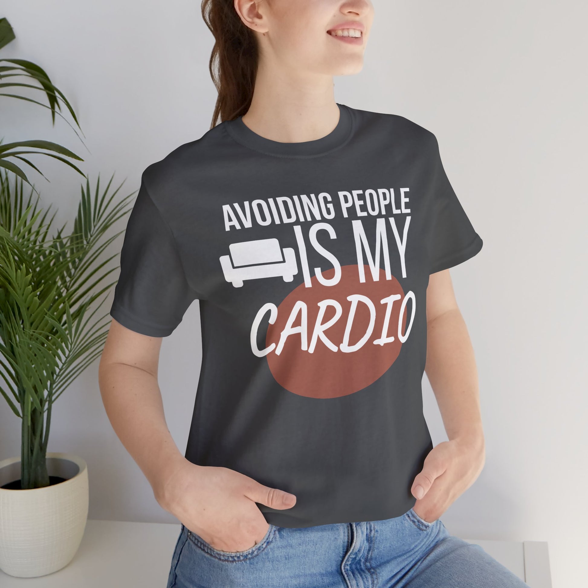 Person wearing a black t-shirt with humorous text 'Avoiding People Is My Cardio' design, standing indoors.