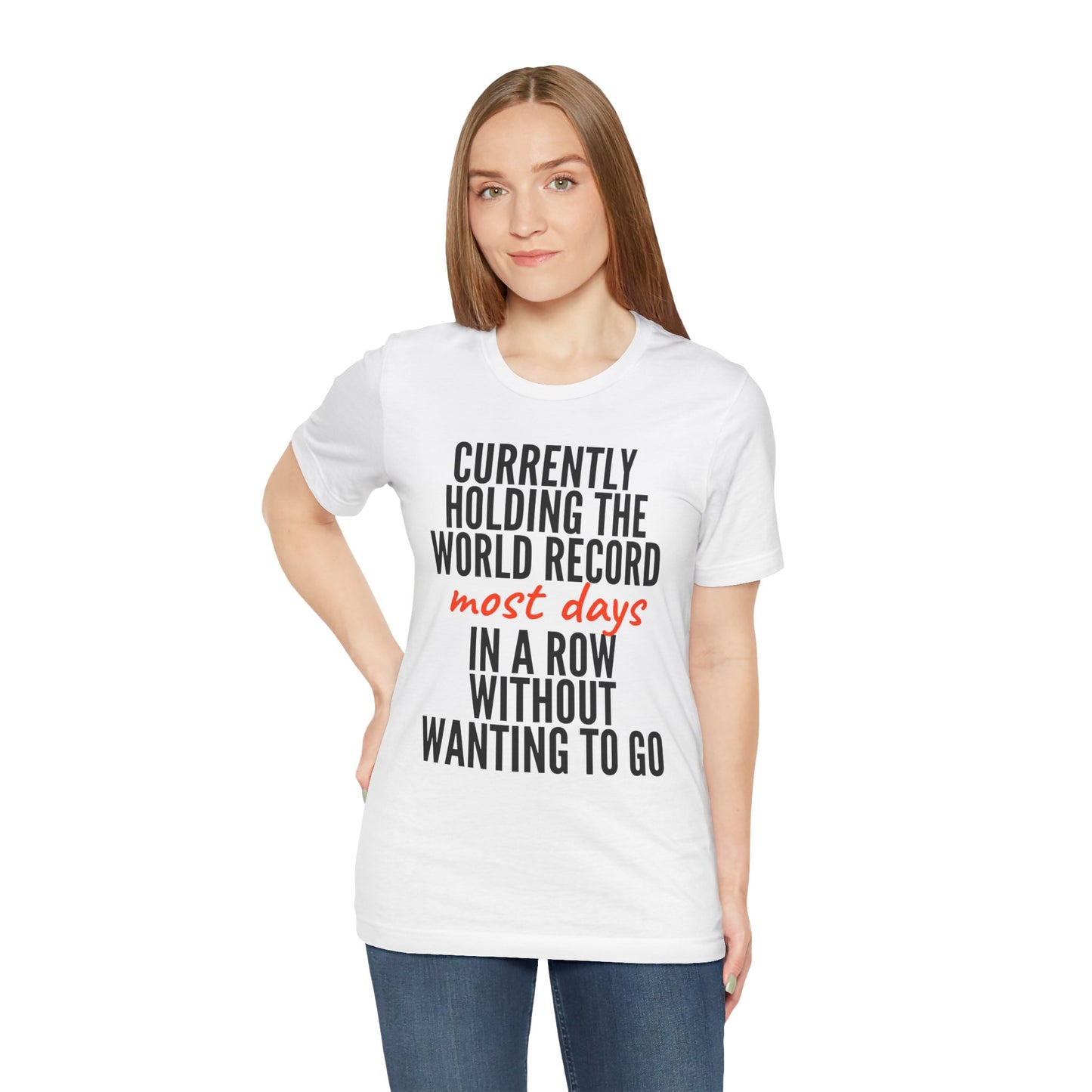 Woman wearing a white t-shirt with humorous text 'Currently holding the world record most days in a row without wanting to go' design on a plain background
