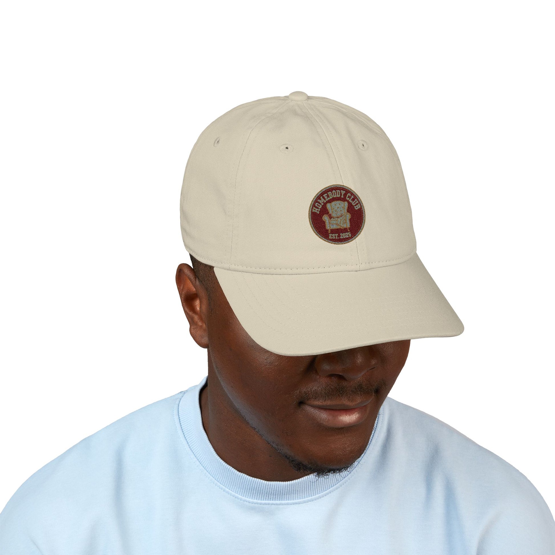 Man wearing a beige cap with a logo and text 'Homebody club Est. 2025' on a white background