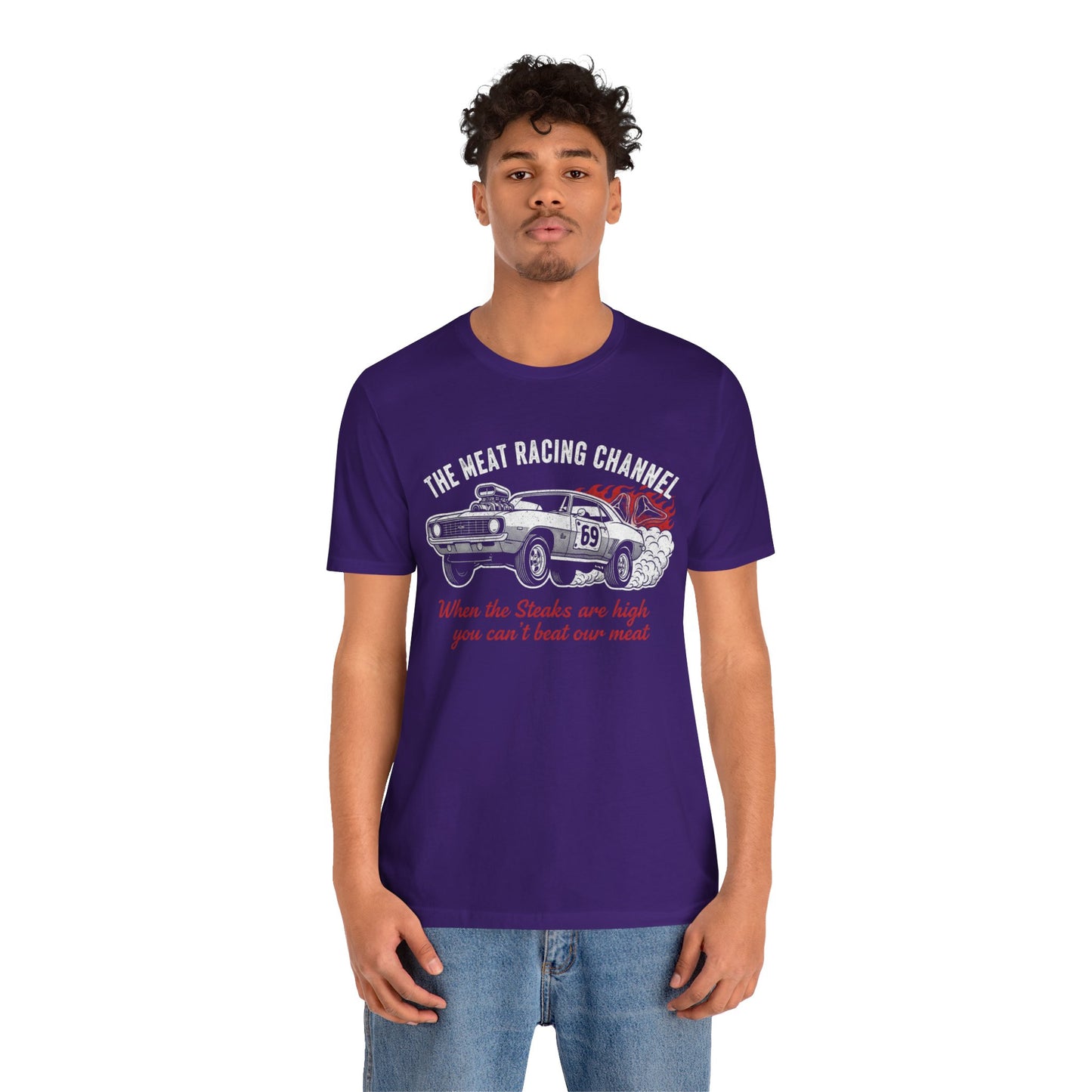 Person wearing a purple Bella+Canvas 3001 T‑shirt featuring a bold drag racing graphic. The design shows a vintage muscle car with the number 69, white smoke and red flames, the distressed white title “The Meat Racing Channel” above, and the red script tagline “When the Steaks are high you can’t beat our meat” below. Funny drag racing parody shirt with retro hot rod style, perfect for car enthusiasts, gearheads, and humor‑driven racing apparel fans.