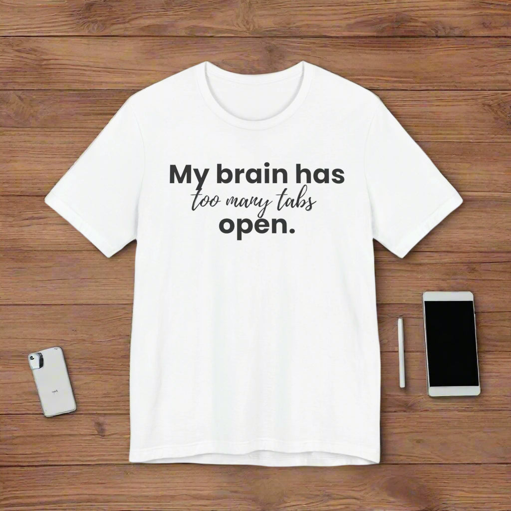 White t-shirt with black text 'My brain has too many tabs open.' on a wood background