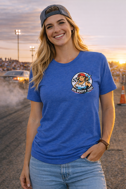 Woman wearing a blue t-shirt with a logo and 'Big Booty Judy' text, standing in an outdoor setting with a sunset.