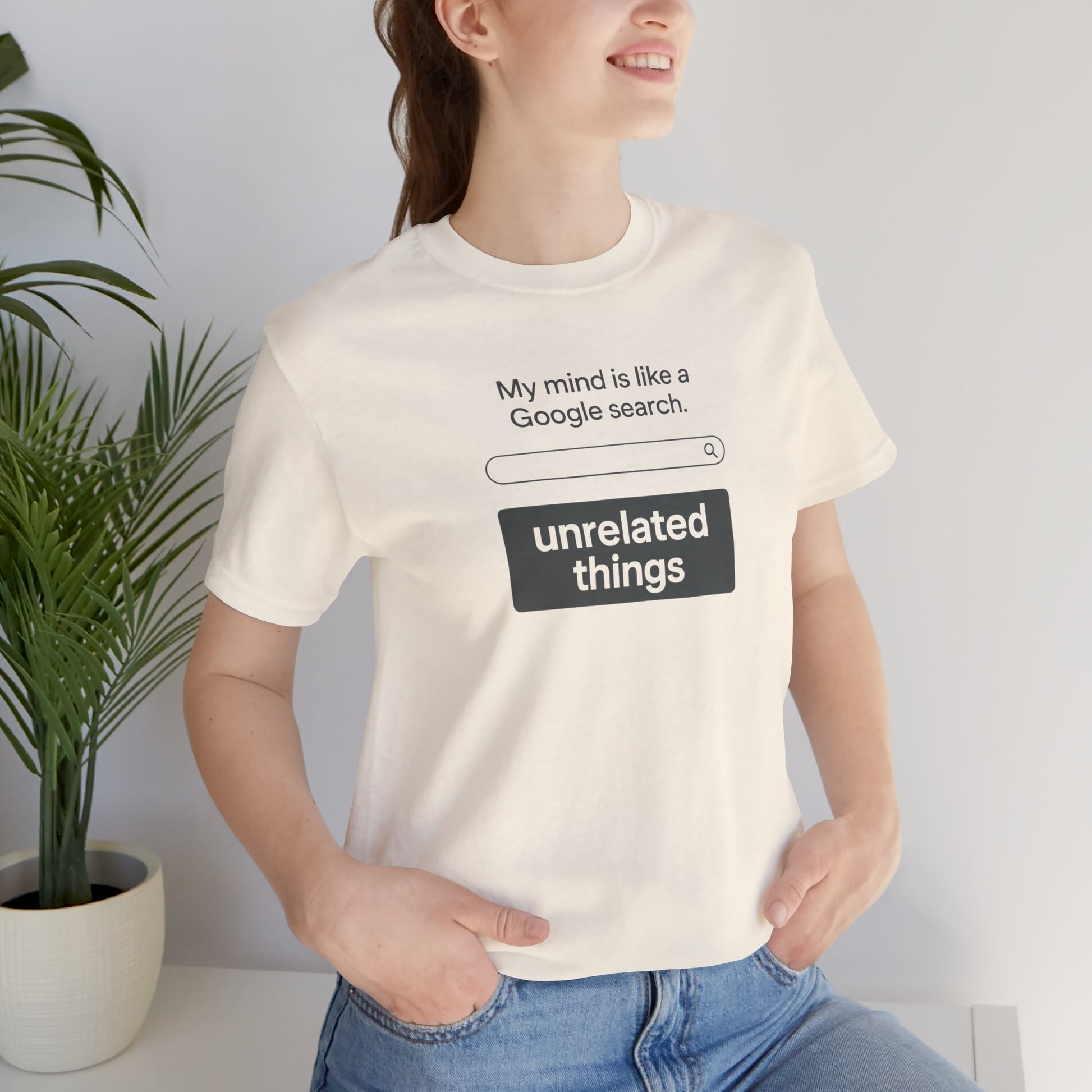 Person wearing a natural t-shirt with humorous text design, standing indoors next to a plant. T-shirt has a search bar graphic with text above that says, 'My mind is like a Google search' and text below that says, 'unrelated things.'