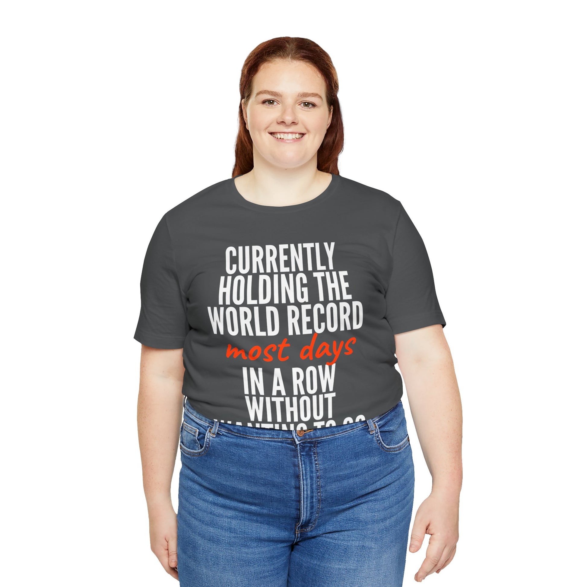 Person wearing a gray t-shirt with humorous text 'Currently holding the world record most days in a row without wanting to go' design on a white background